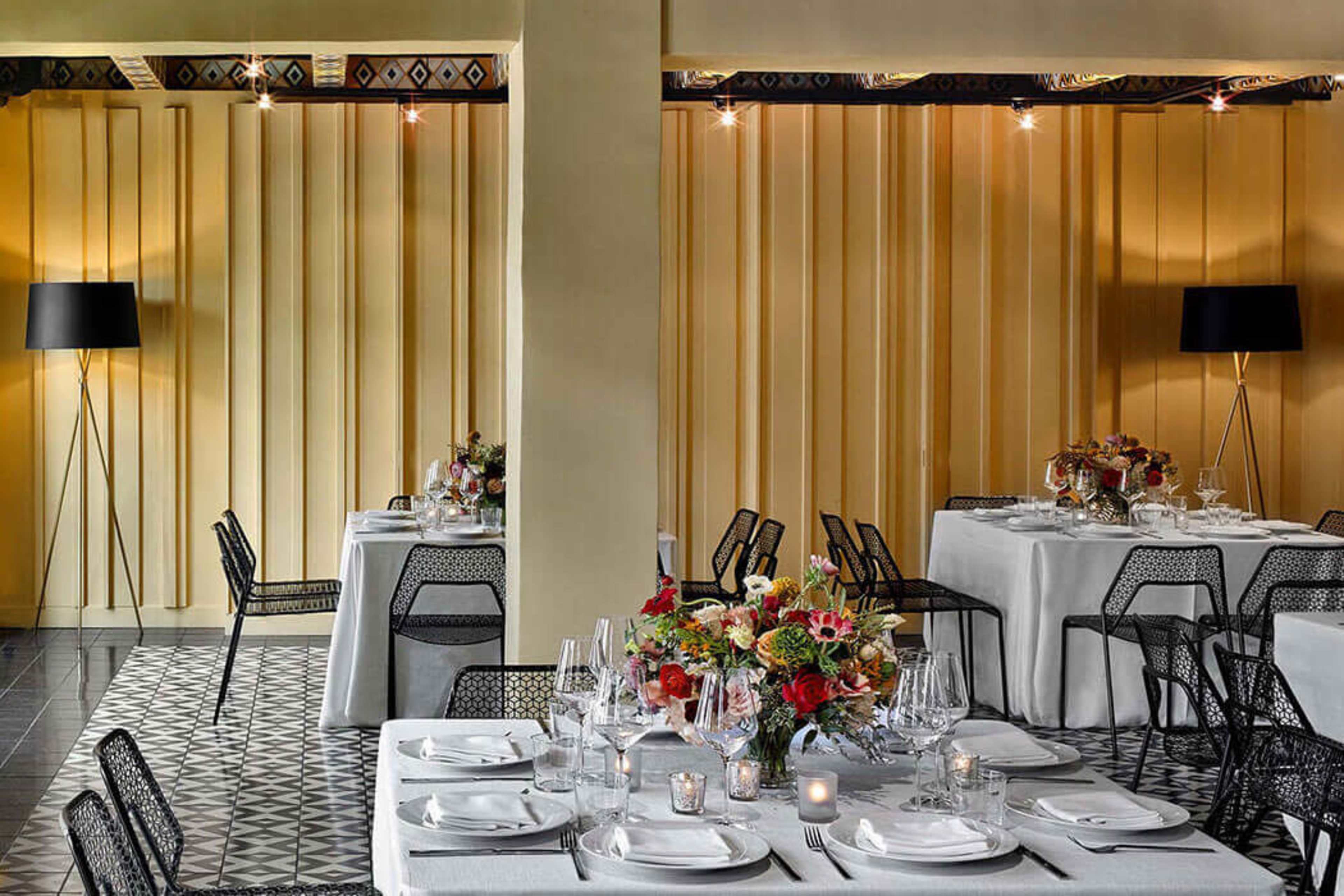 The image shows a modern dining space with elegantly set tables featuring white tablecloths, glassware, and floral centerpieces, complemented by a yellow accent wall and stylish black chairs.