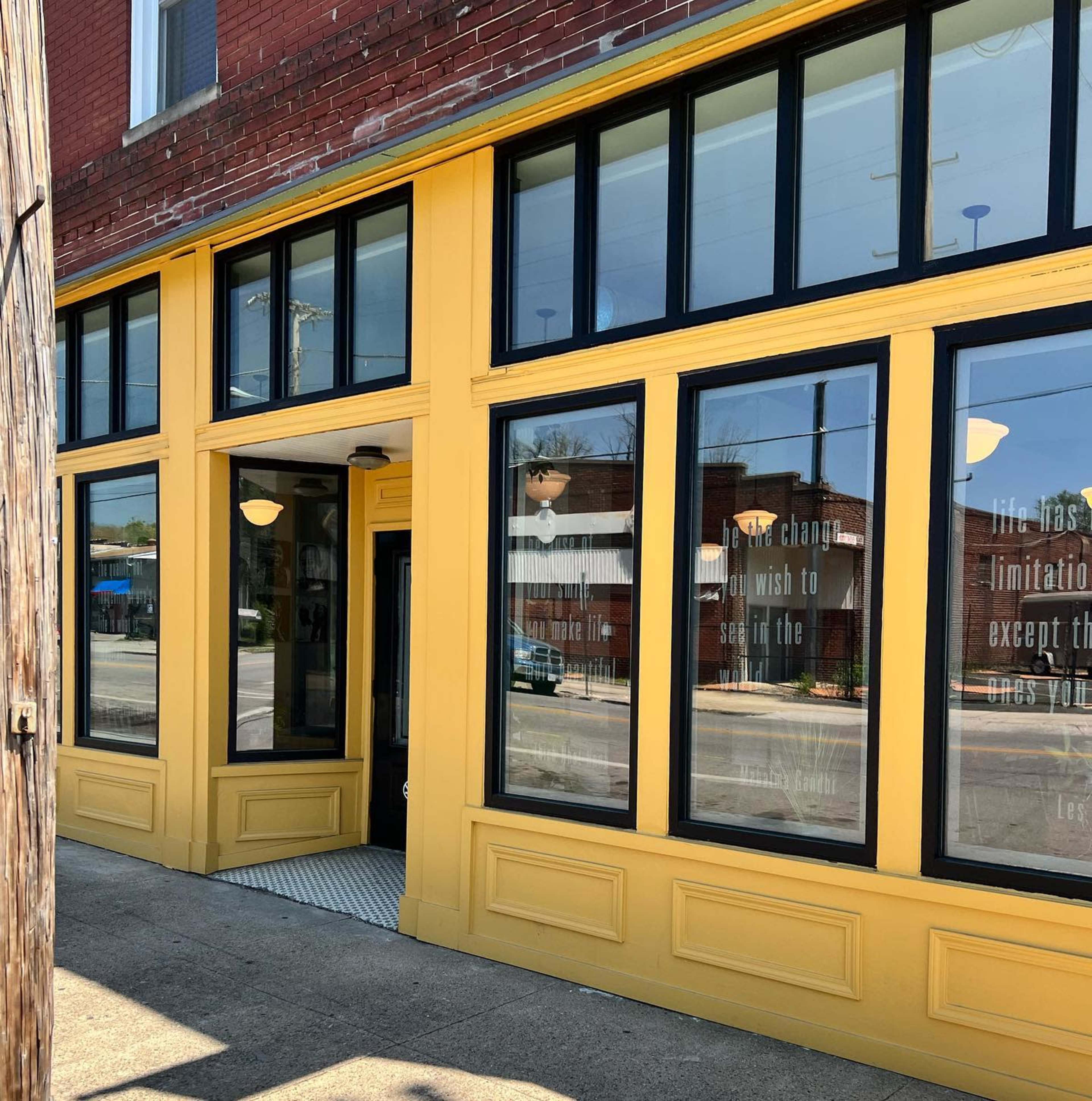 A storefront with large windows and a yellow exterior features motivational quotes on the glass, located on a street lined with brick buildings.