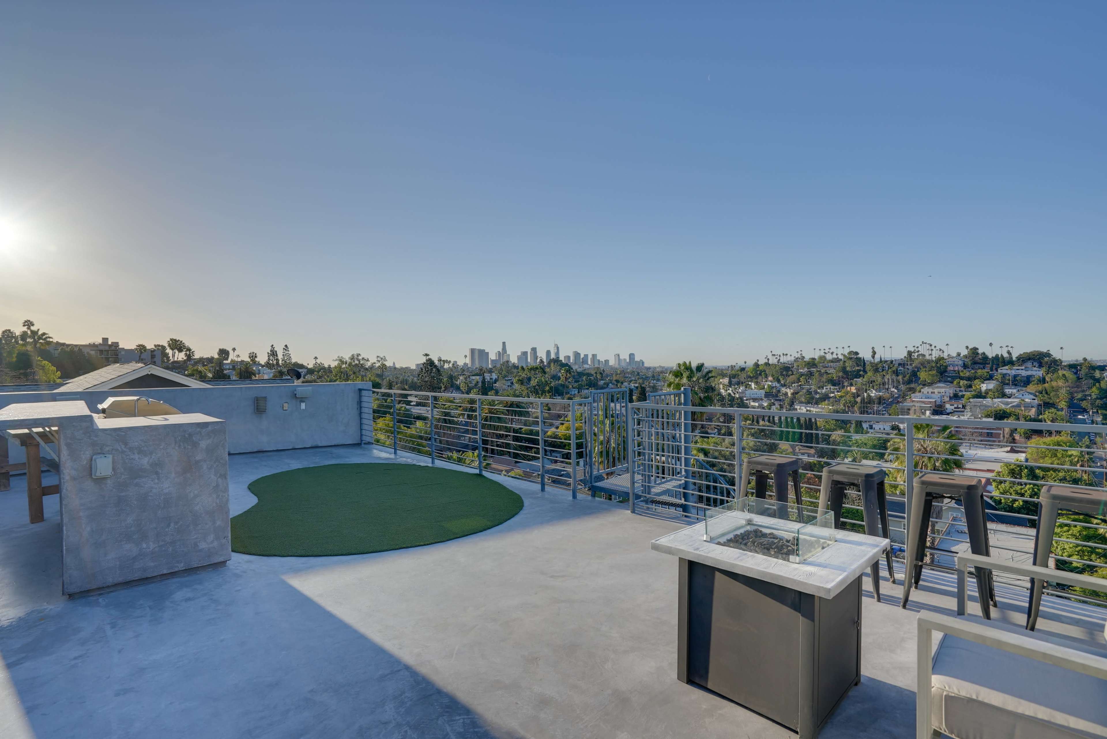 A rooftop patio featuring a green turf area, a fire pit, and a view of the city skyline in the distance.