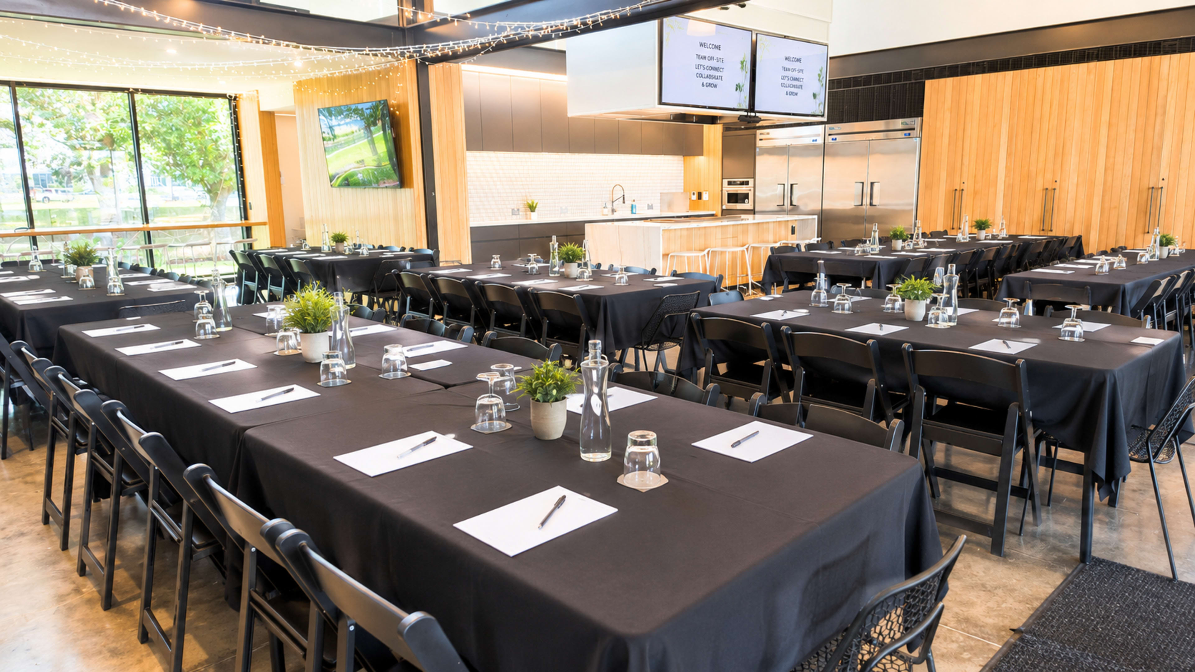 The image shows a large, modern conference room arranged with several long tables covered in black tablecloths, each set with glassware and notepads, and featuring windows that allow natural light to fill the space.