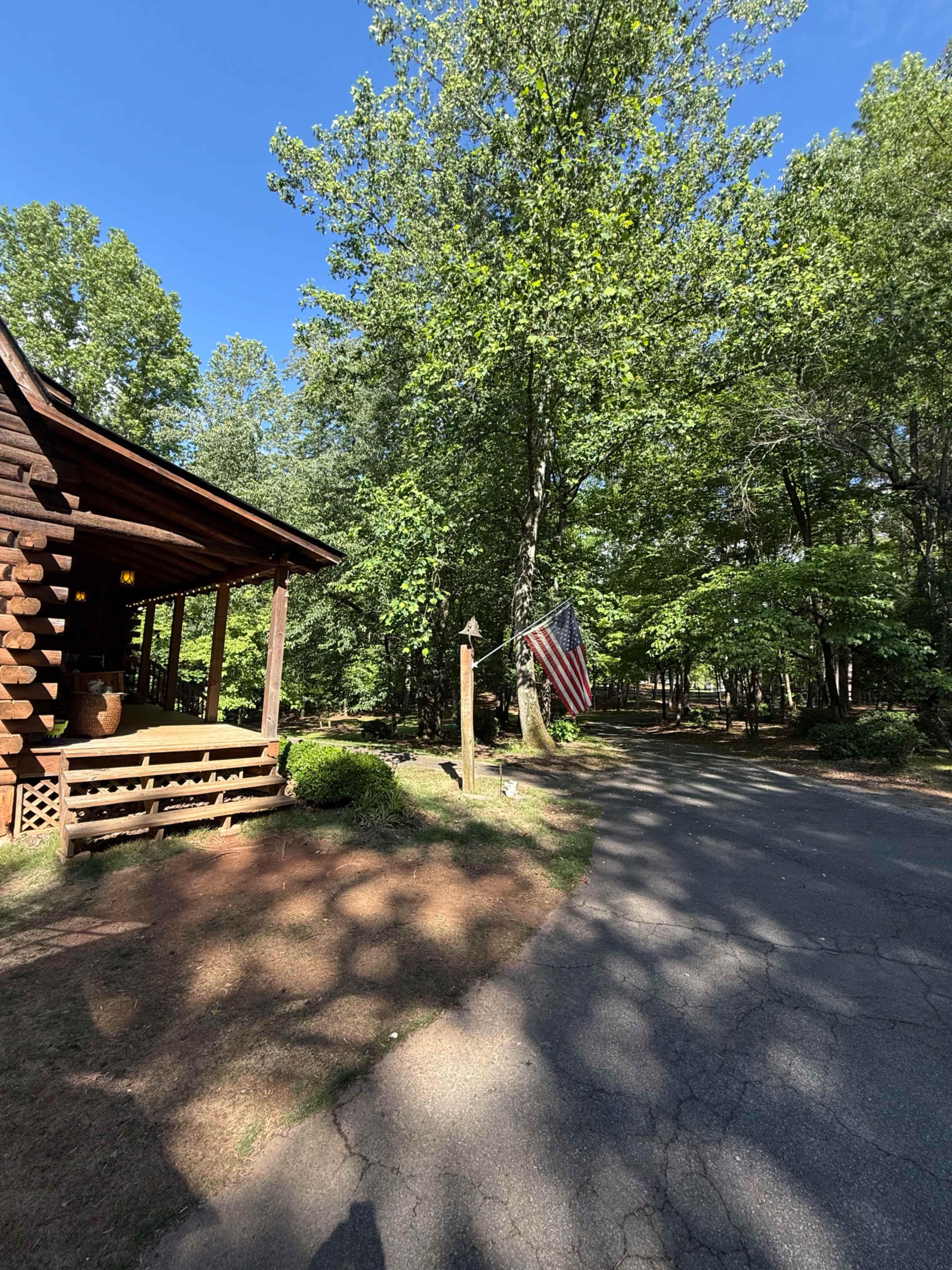A log cabin with a porch and an American flag hangs from a post beside a gravel driveway surrounded by trees.