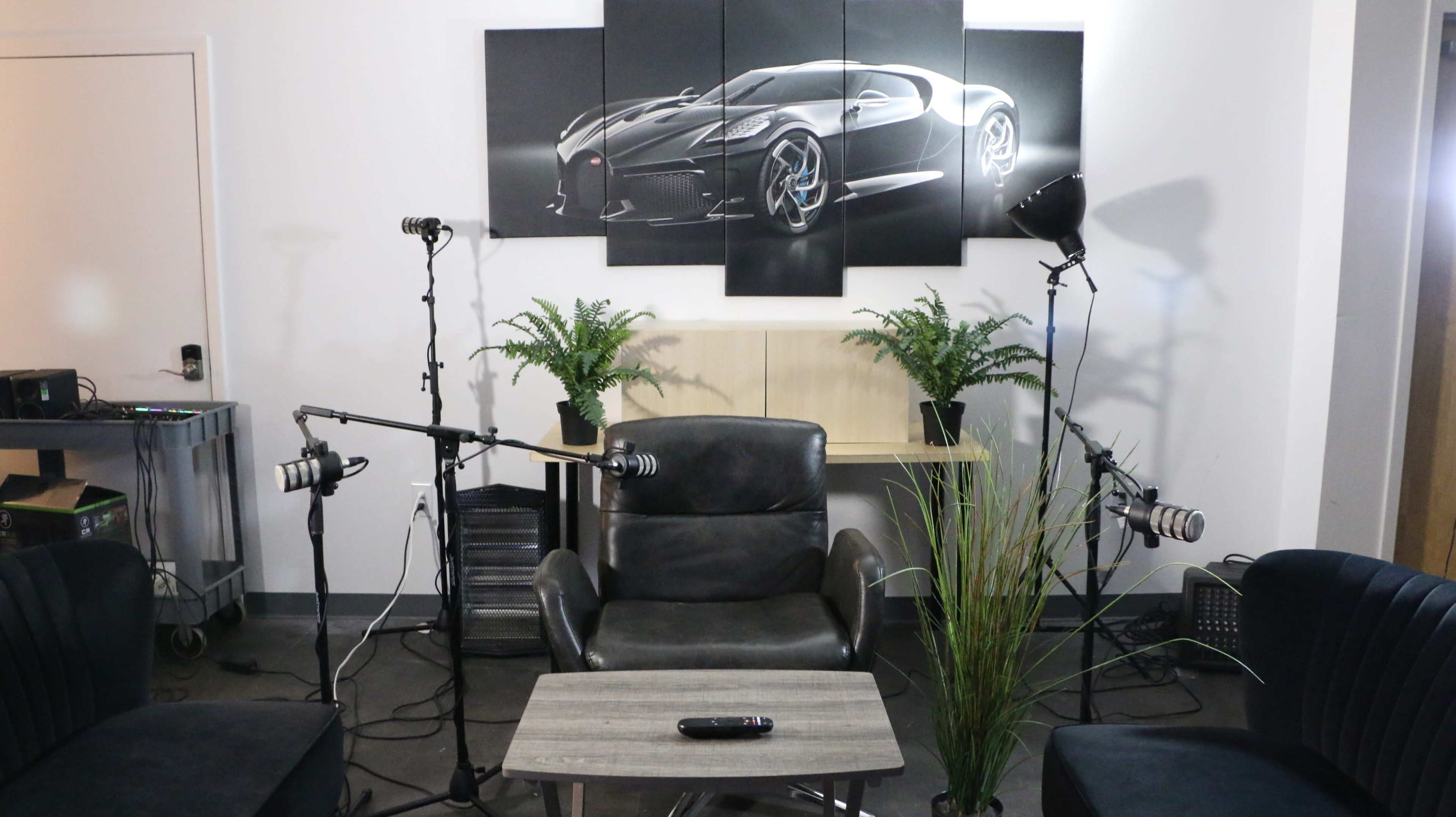 The image shows a modern interview setup featuring a central chair, a wooden table, and several microphones positioned around it, with a large automotive artwork on the wall.