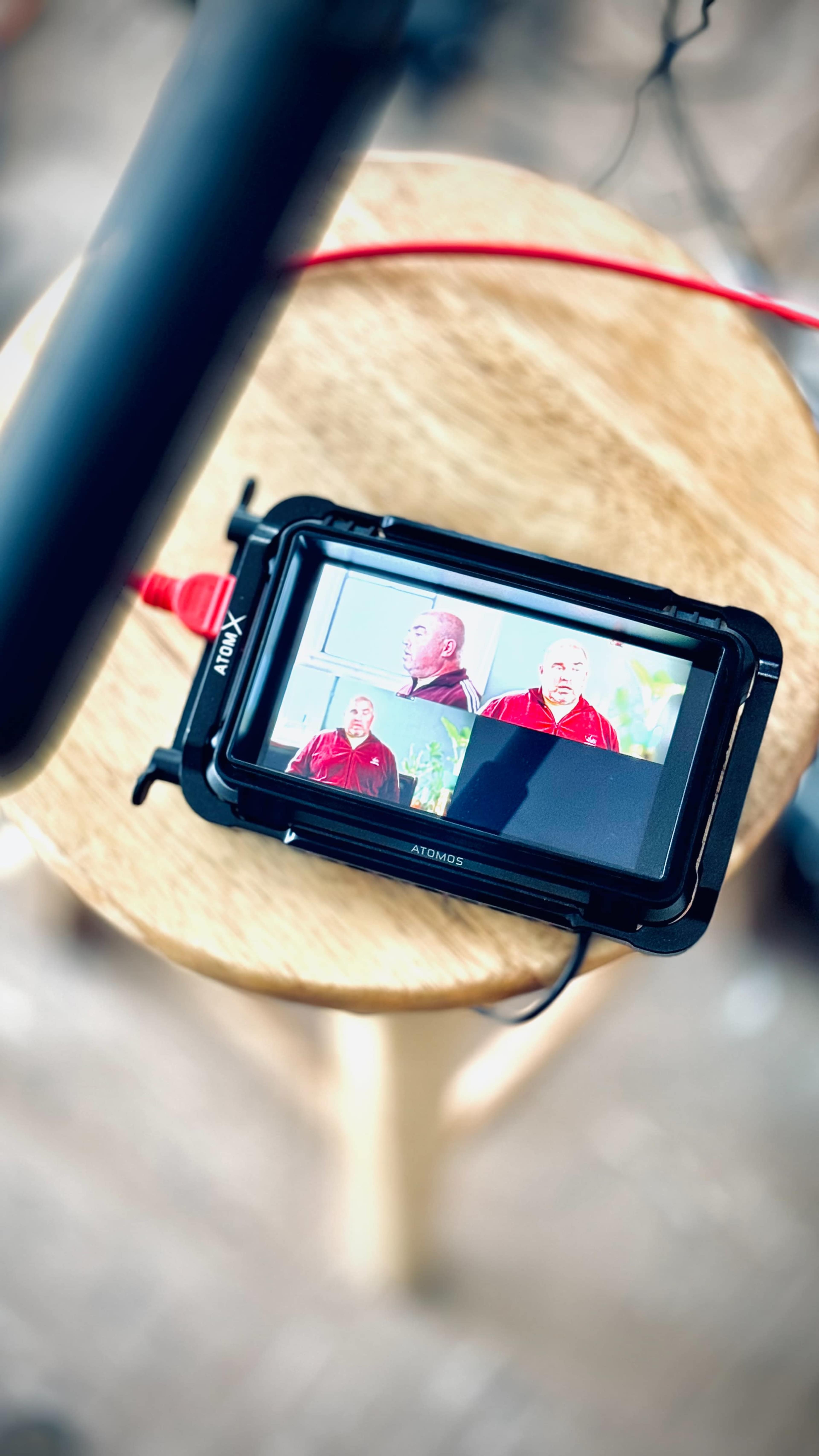 A camera monitor displays a man in a red shirt speaking, positioned on a wooden stool.