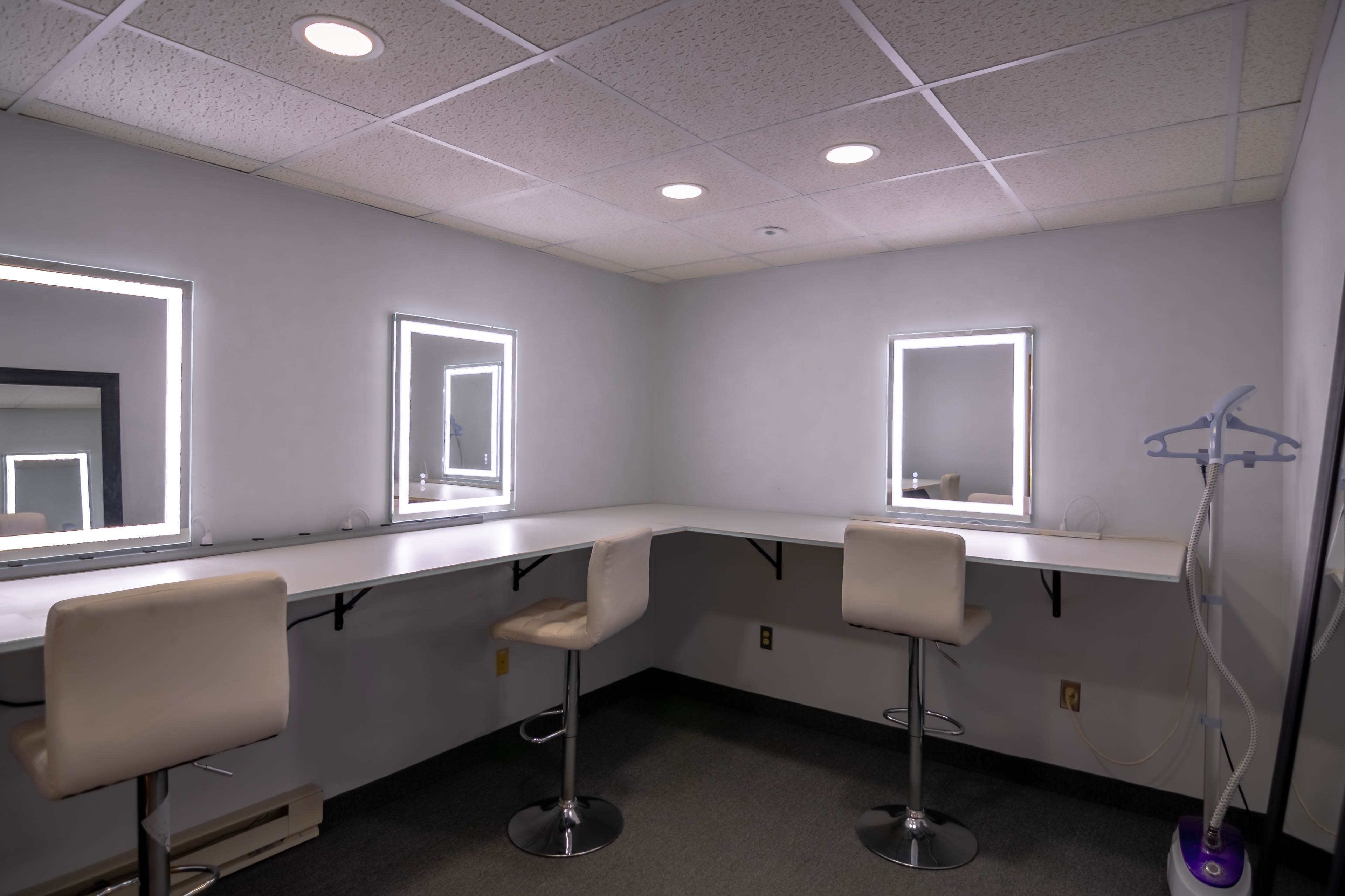 The image shows a brightly lit dressing room with two illuminated mirrors and two stools along a white countertop.