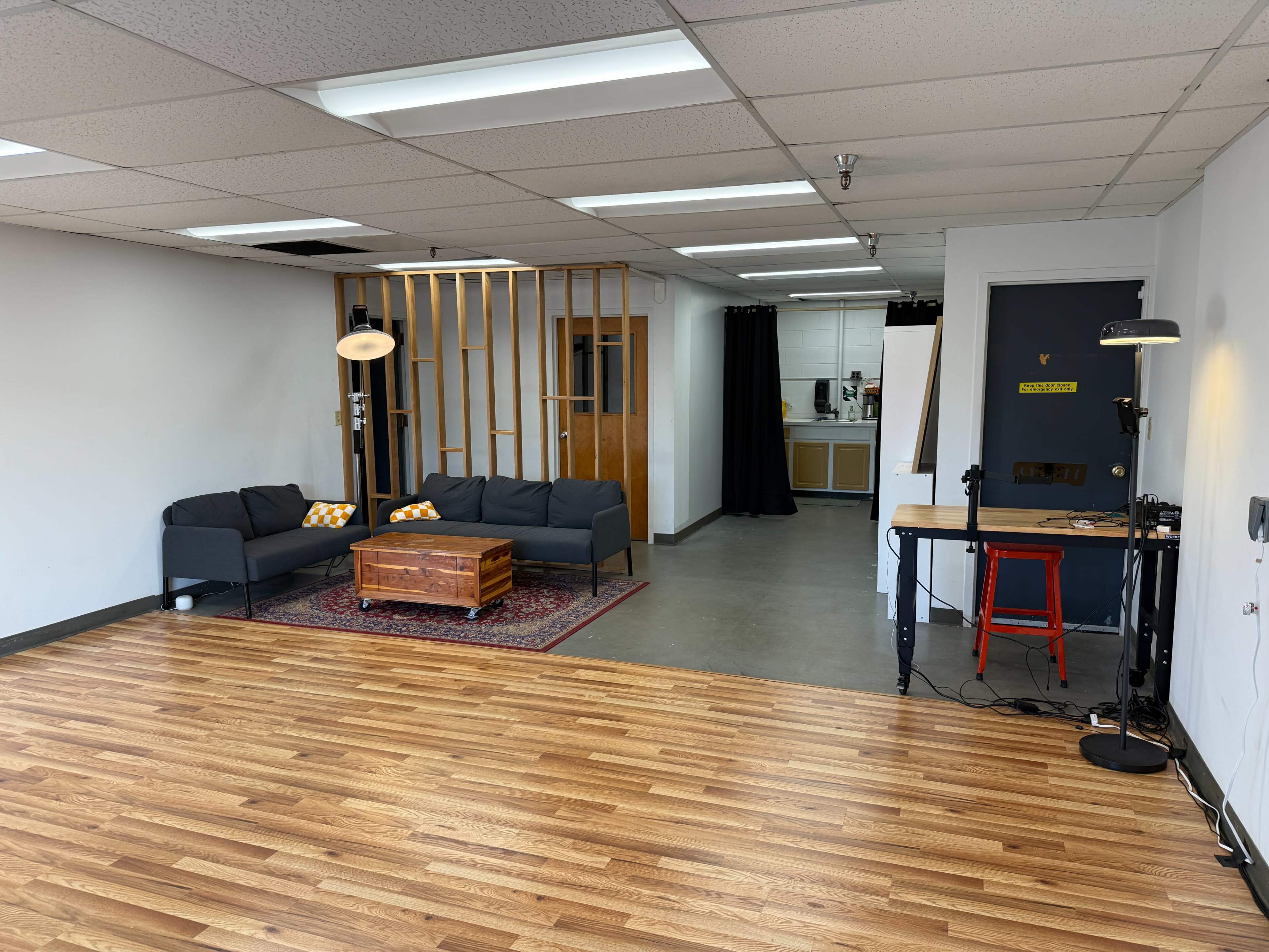 The image shows a sparsely furnished interior space with a black couch, a wooden coffee table, and a small area with a light and a desk, leading into a kitchen area in the background.