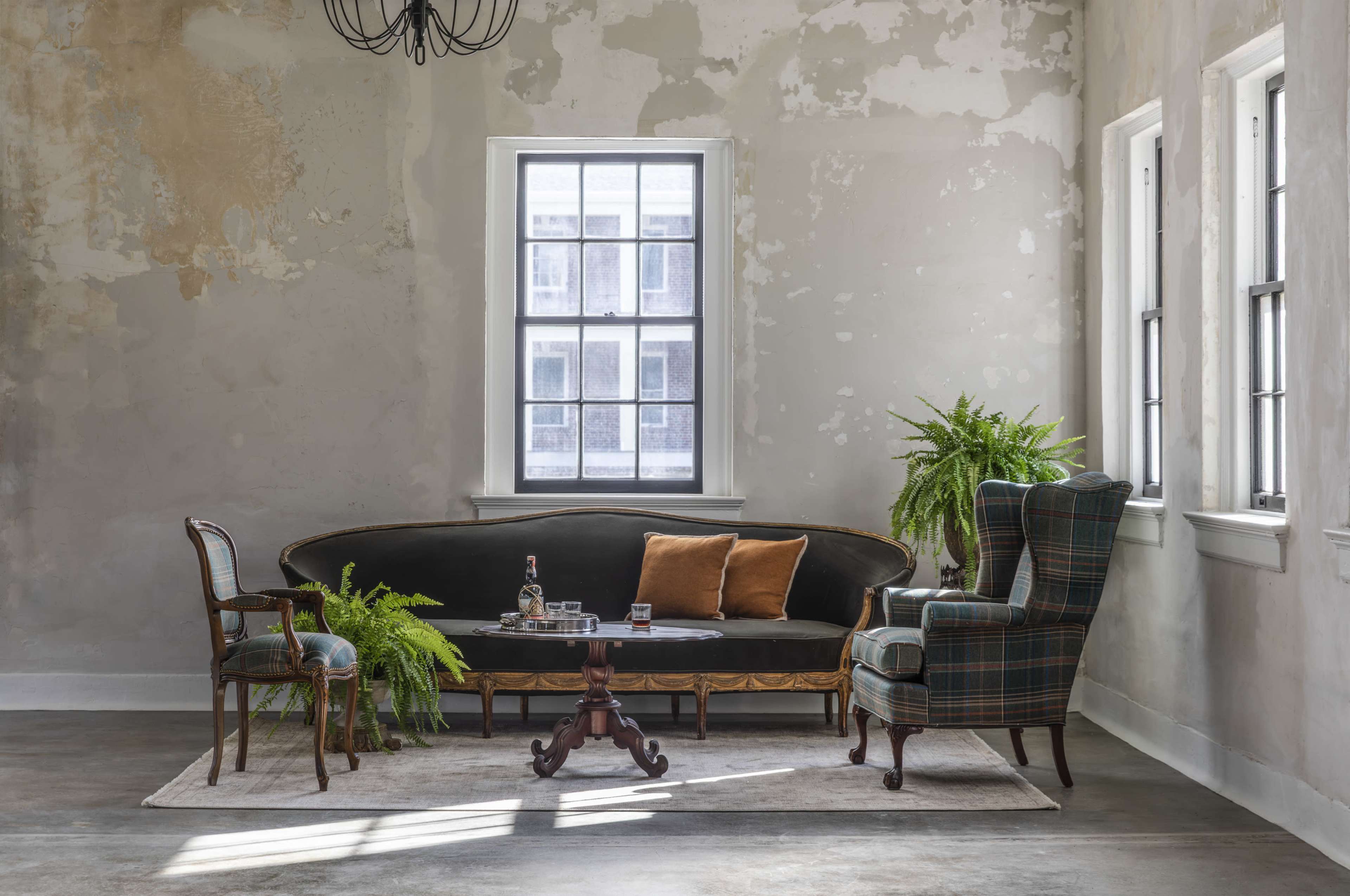 A vintage sofa and chairs are arranged in a room with peeling walls and large windows, illuminated by natural light, alongside potted ferns and a small table.
