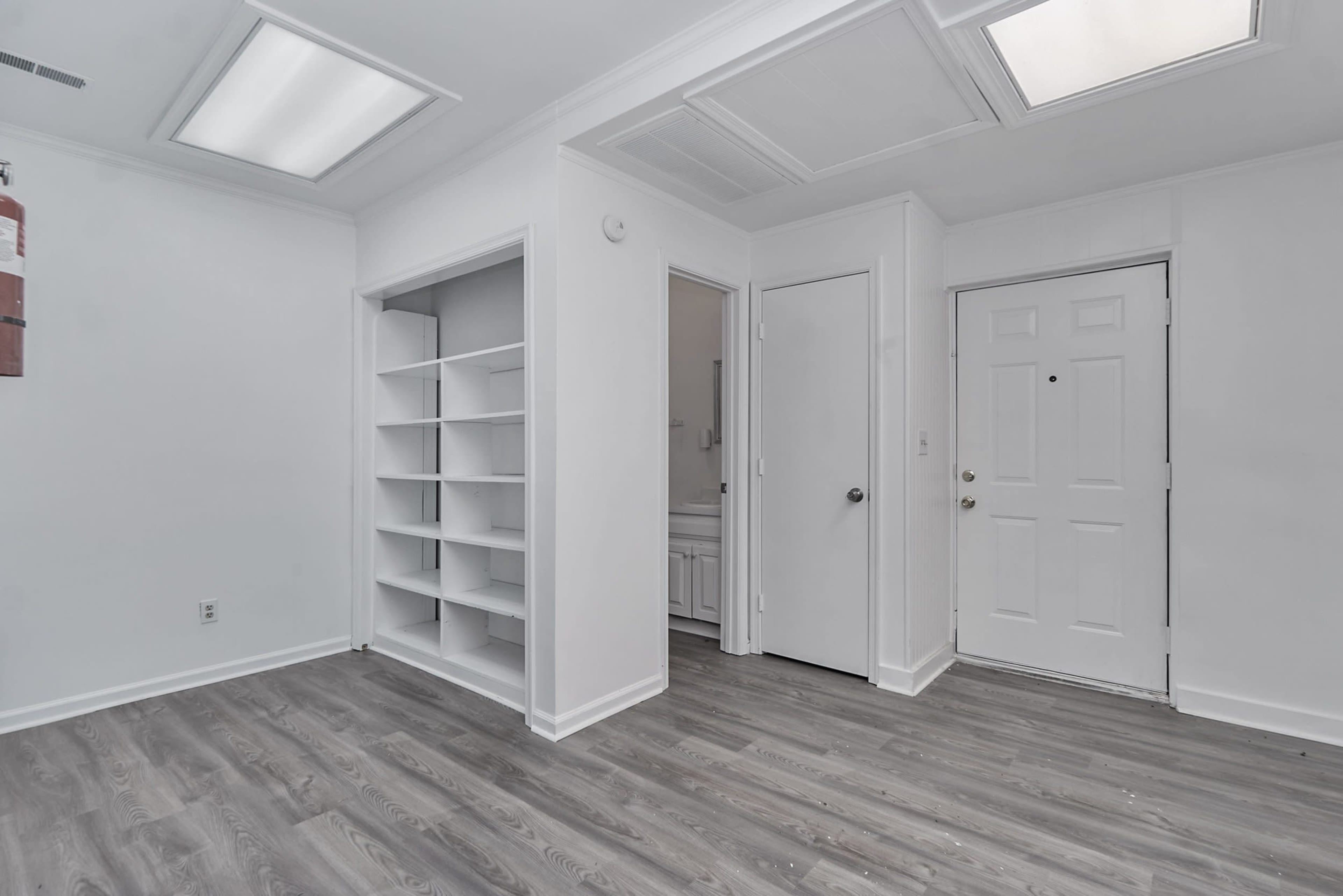 The image shows a minimalist interior space with a white wall, a door on the right, and a built-in bookshelf on the left.