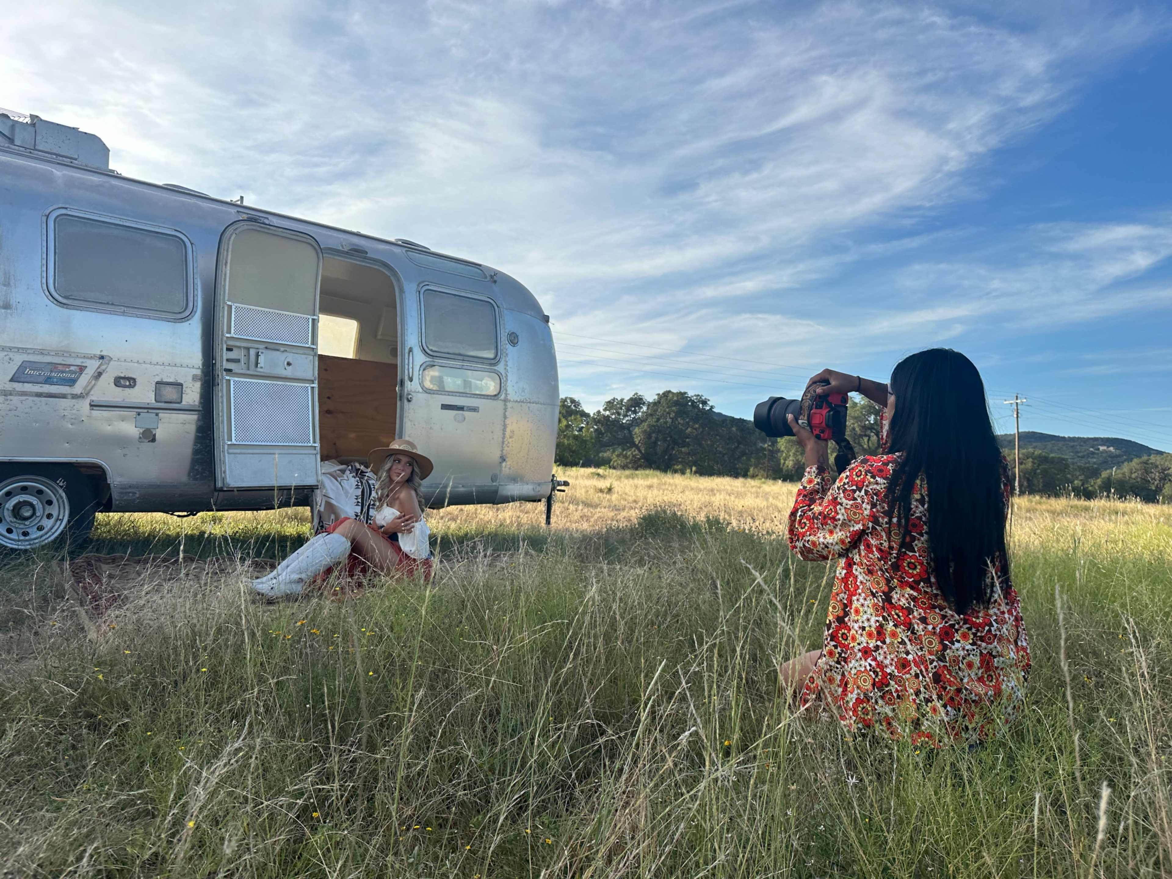 A woman in a floral dress photographs another woman sitting in an open vintage Airstream trailer on a grassy field.