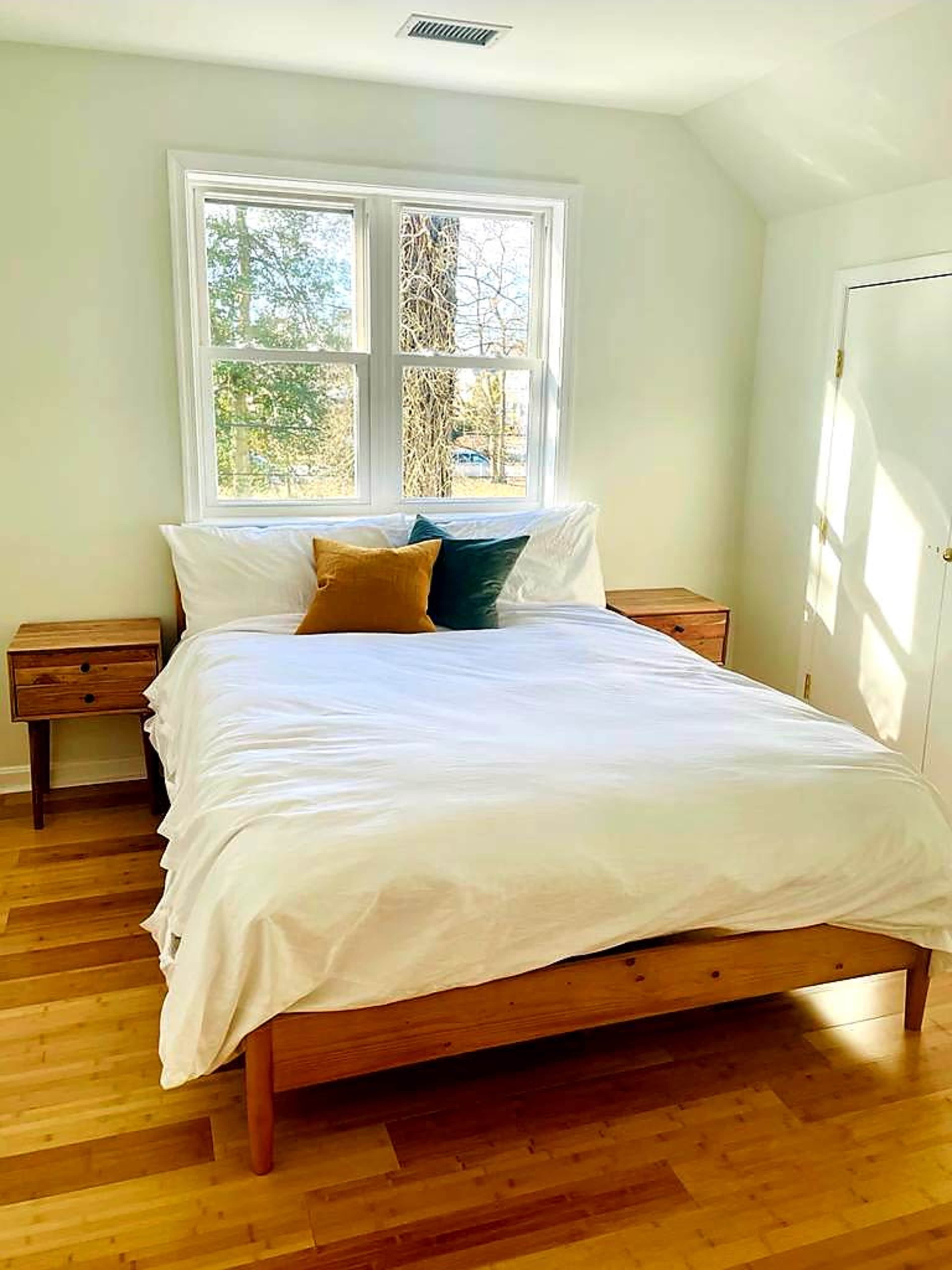 A simple bedroom features a wooden bed with white bedding, two wooden nightstands, and large windows letting in natural light.