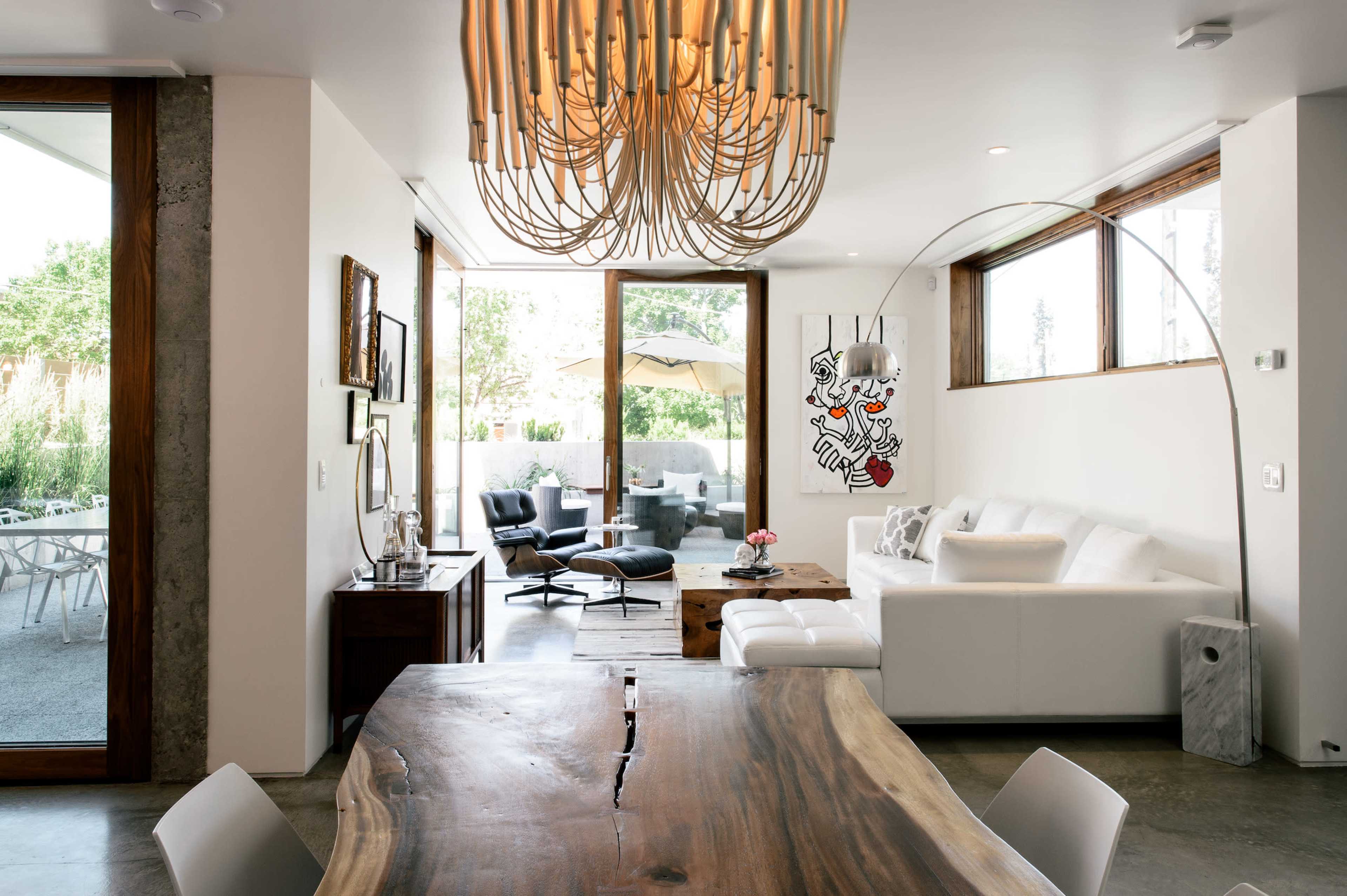The image shows a modern living space featuring a wooden dining table, a white sectional sofa, large windows, and a contemporary chandelier.