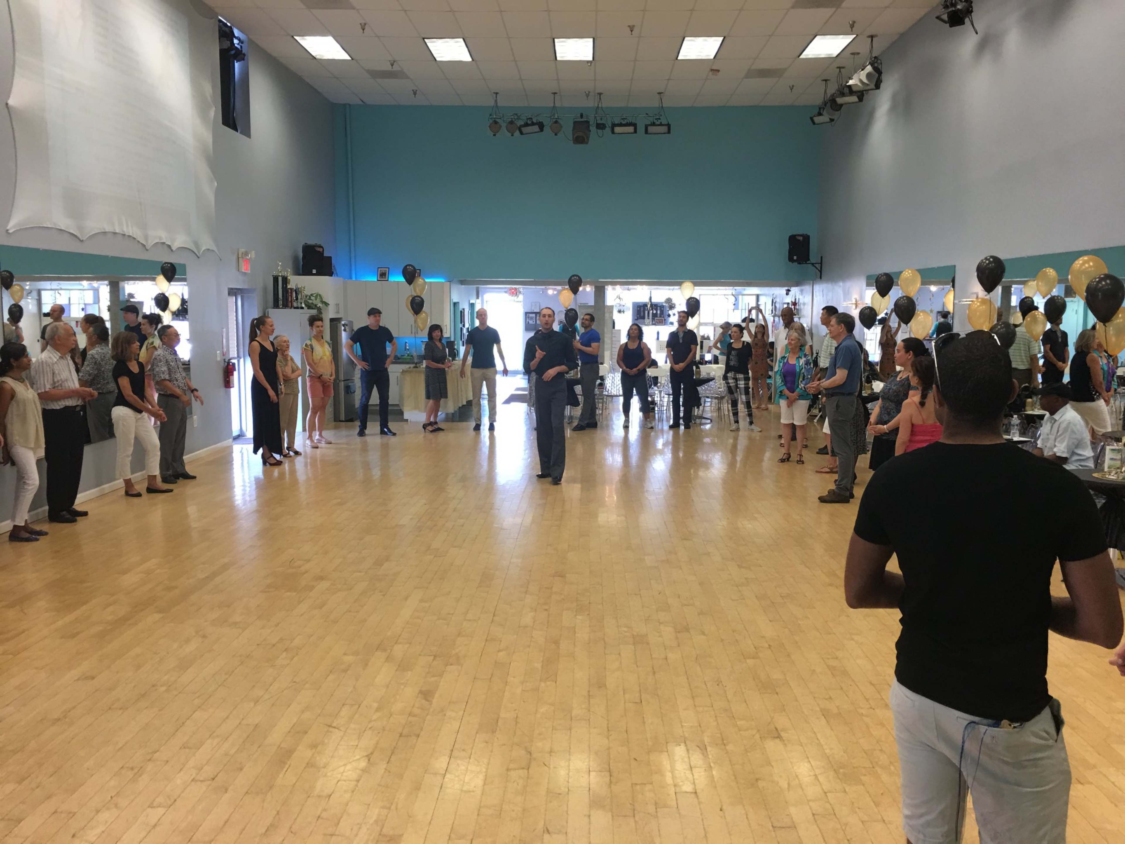 A large group of people stands in a circle inside a spacious dance studio, while a speaker addresses them from the center.