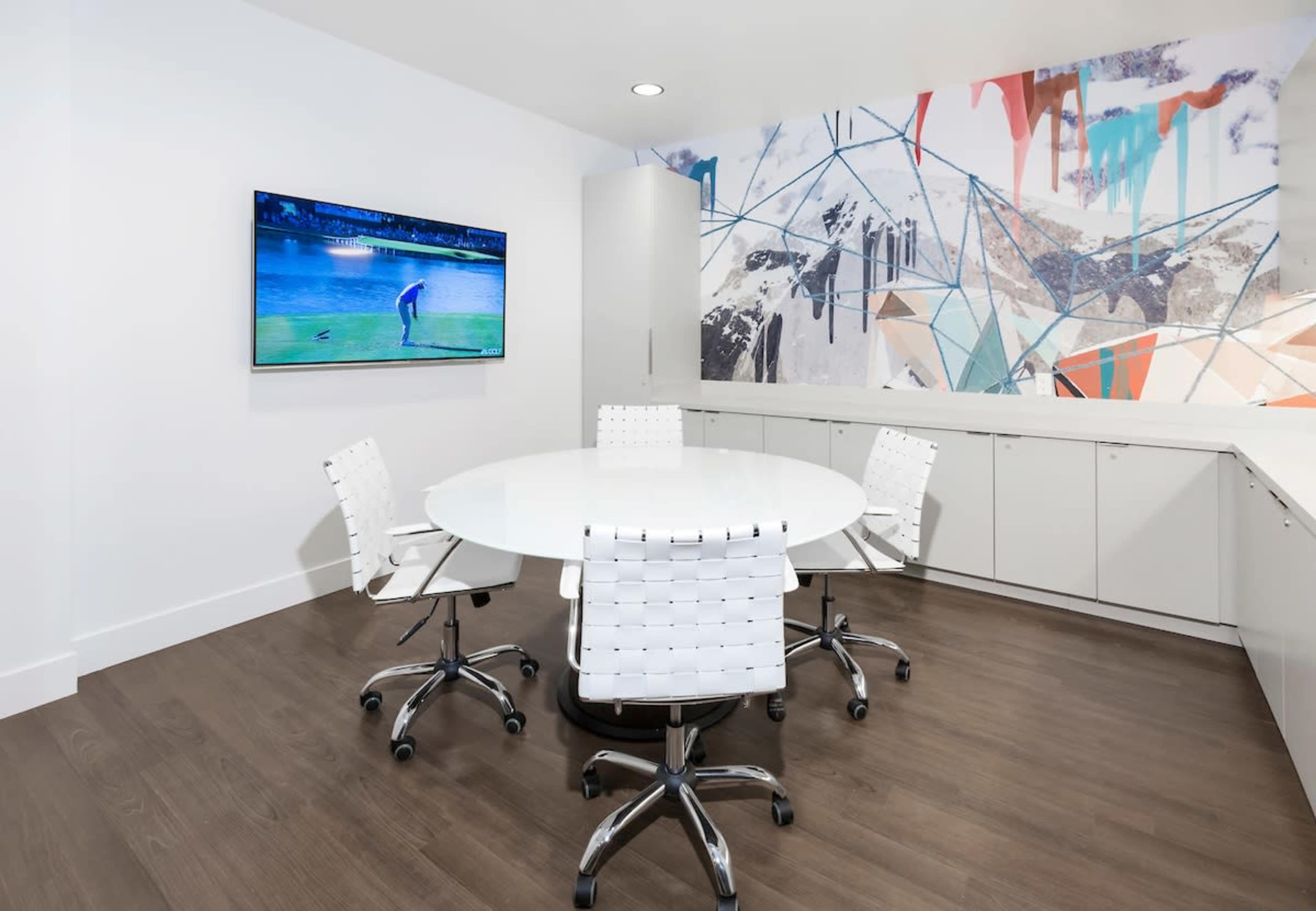 The image shows a modern conference room with a circular table, four white chairs, a television on the wall displaying a golf scene, and a colorful abstract mural.
