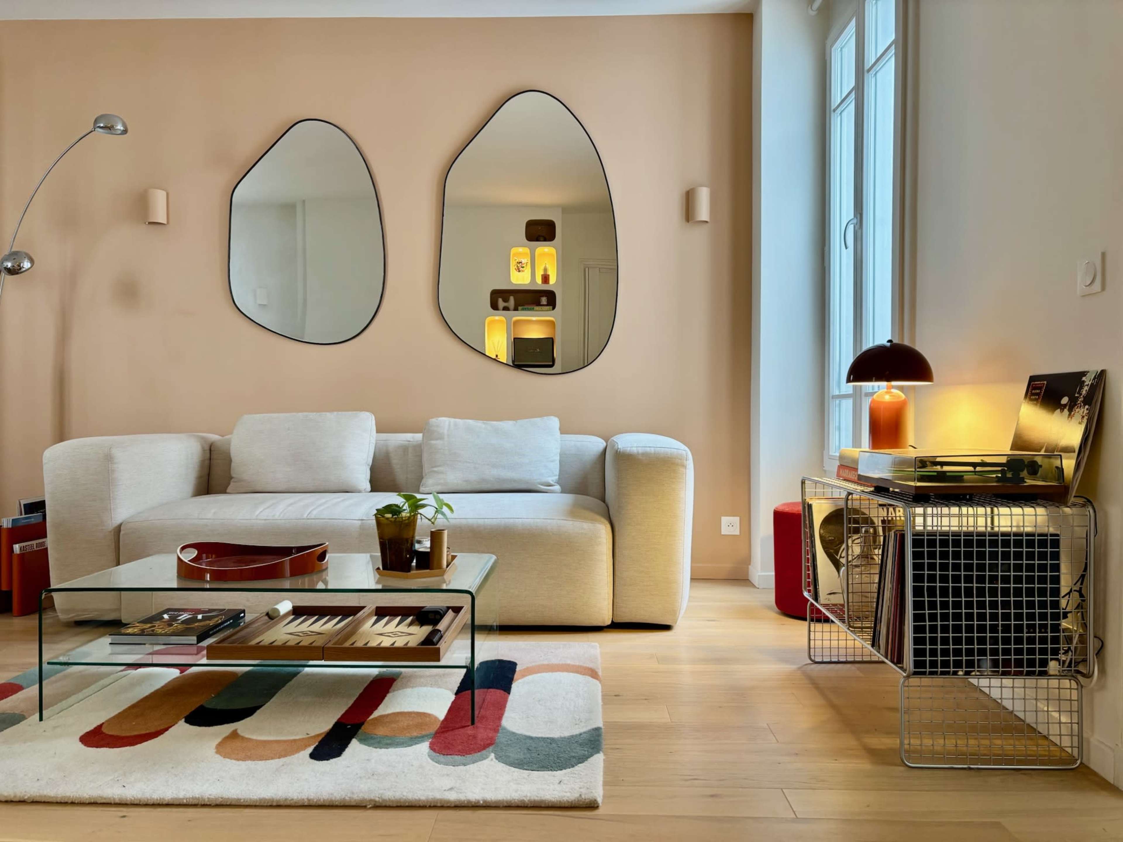 The image shows a modern living room featuring a light-colored sofa, a glass coffee table with books and a plant, a colorful rug, two abstract mirrors on the wall, and a record player with vinyl records on a wire cart.