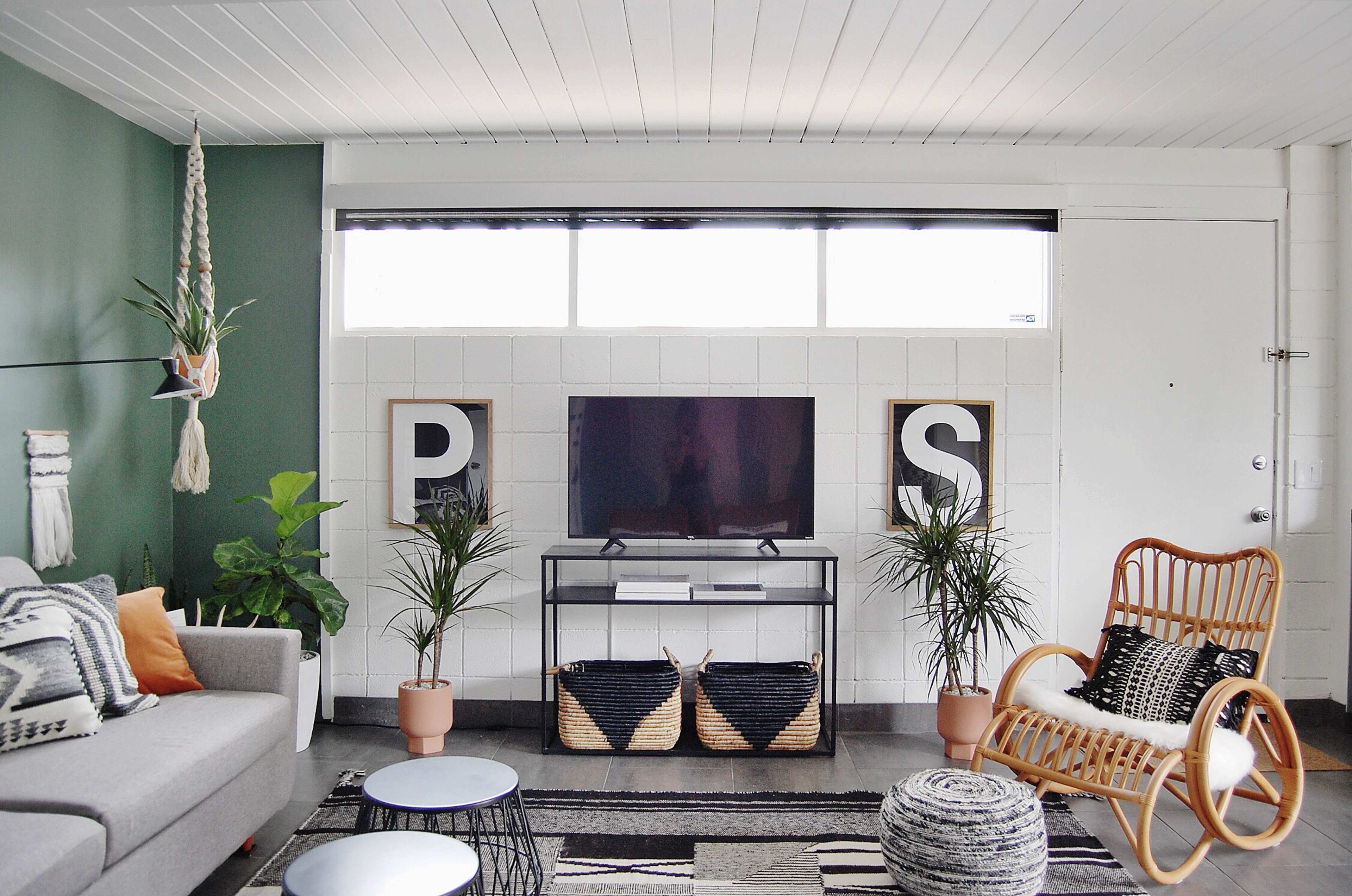 A modern living room features a gray sofa, a television on a metal stand, and decorative plants against a backdrop of green walls and large windows.
