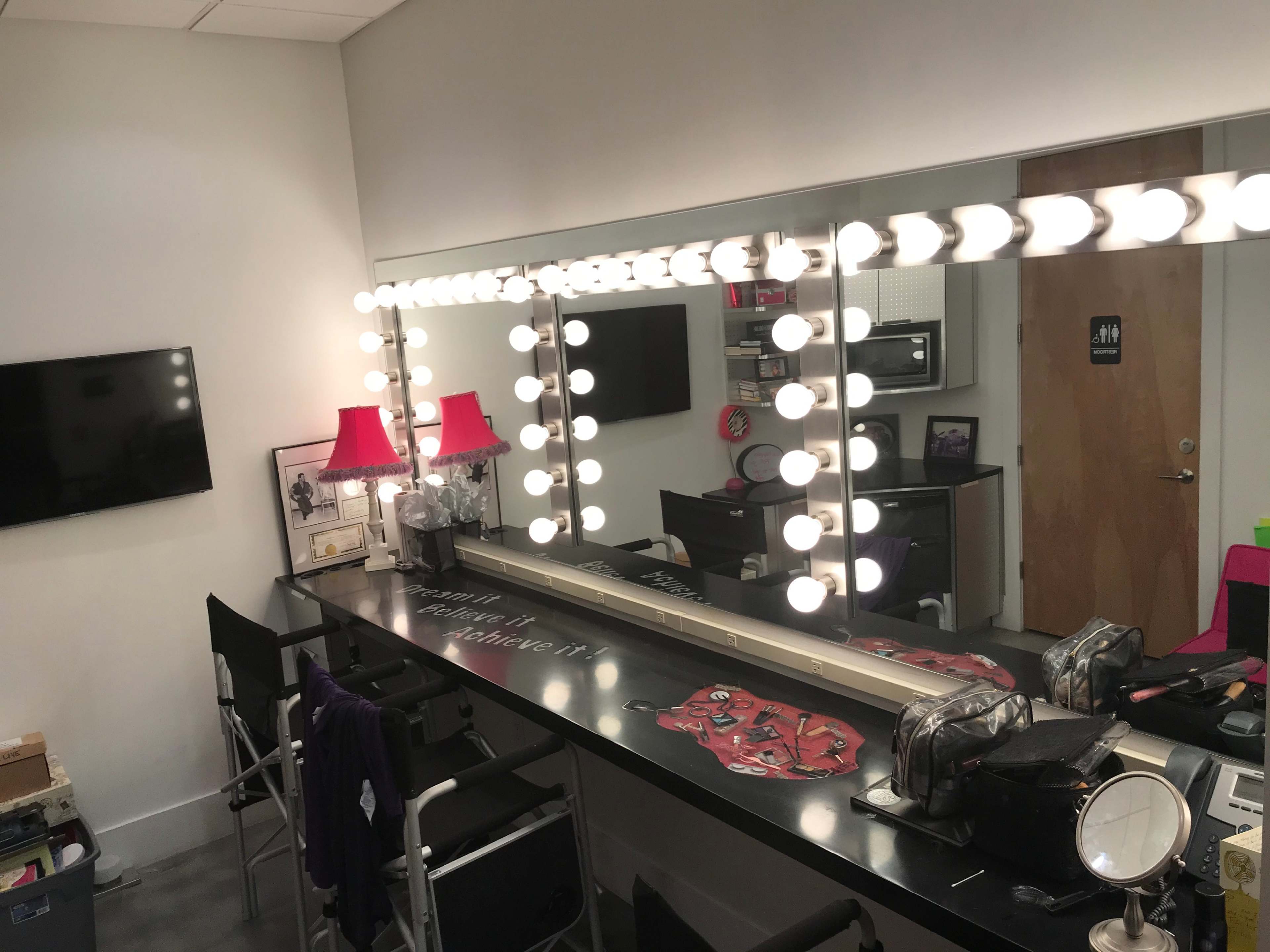 A brightly lit dressing room with a long mirror surrounded by light bulbs, a table with personal items, and a door leading to a restroom.