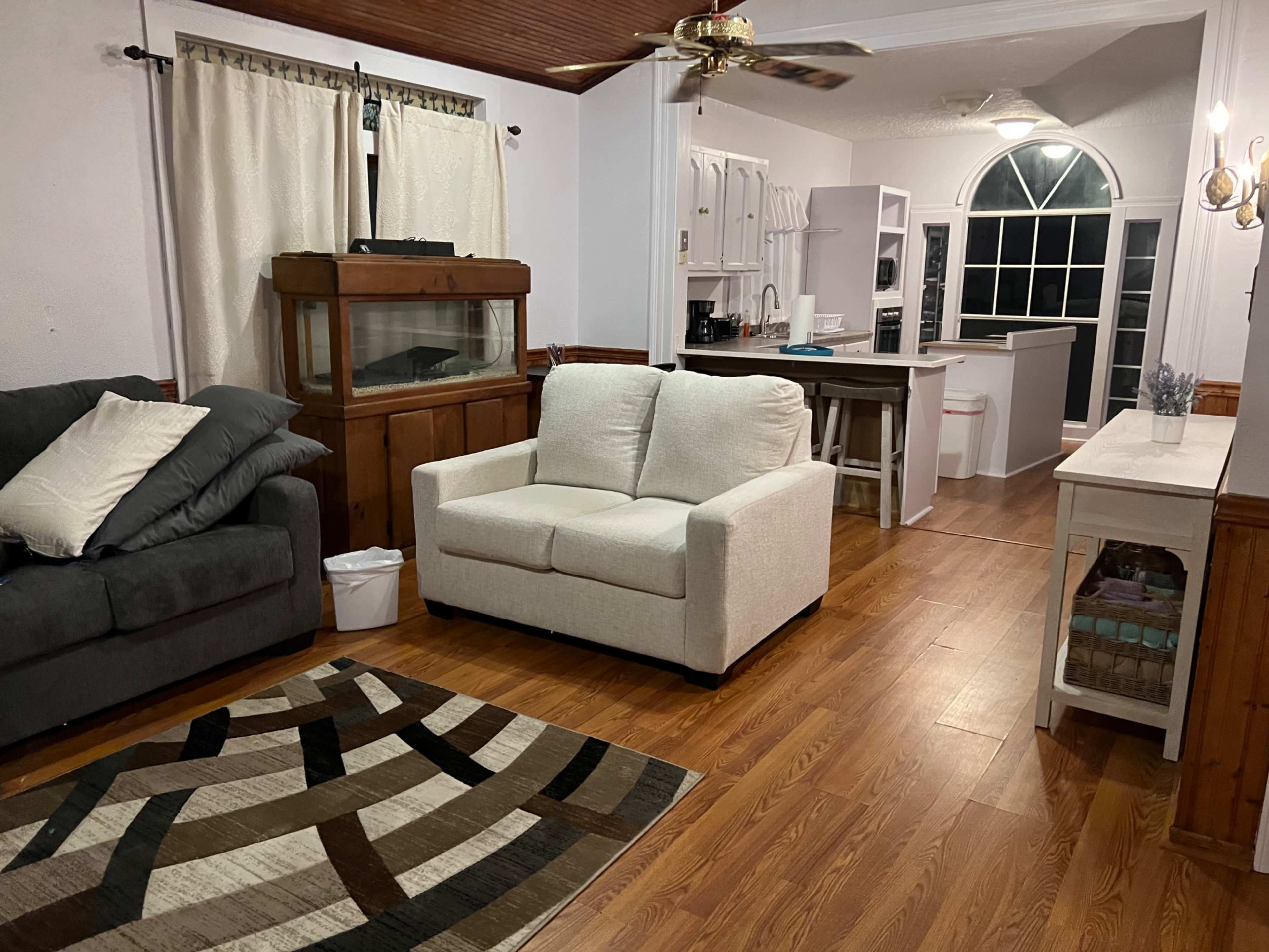 The image shows a living room with a gray couch, a white loveseat, a large rug, and a wood cabinet, leading to a kitchen area with white cabinetry and a large window.