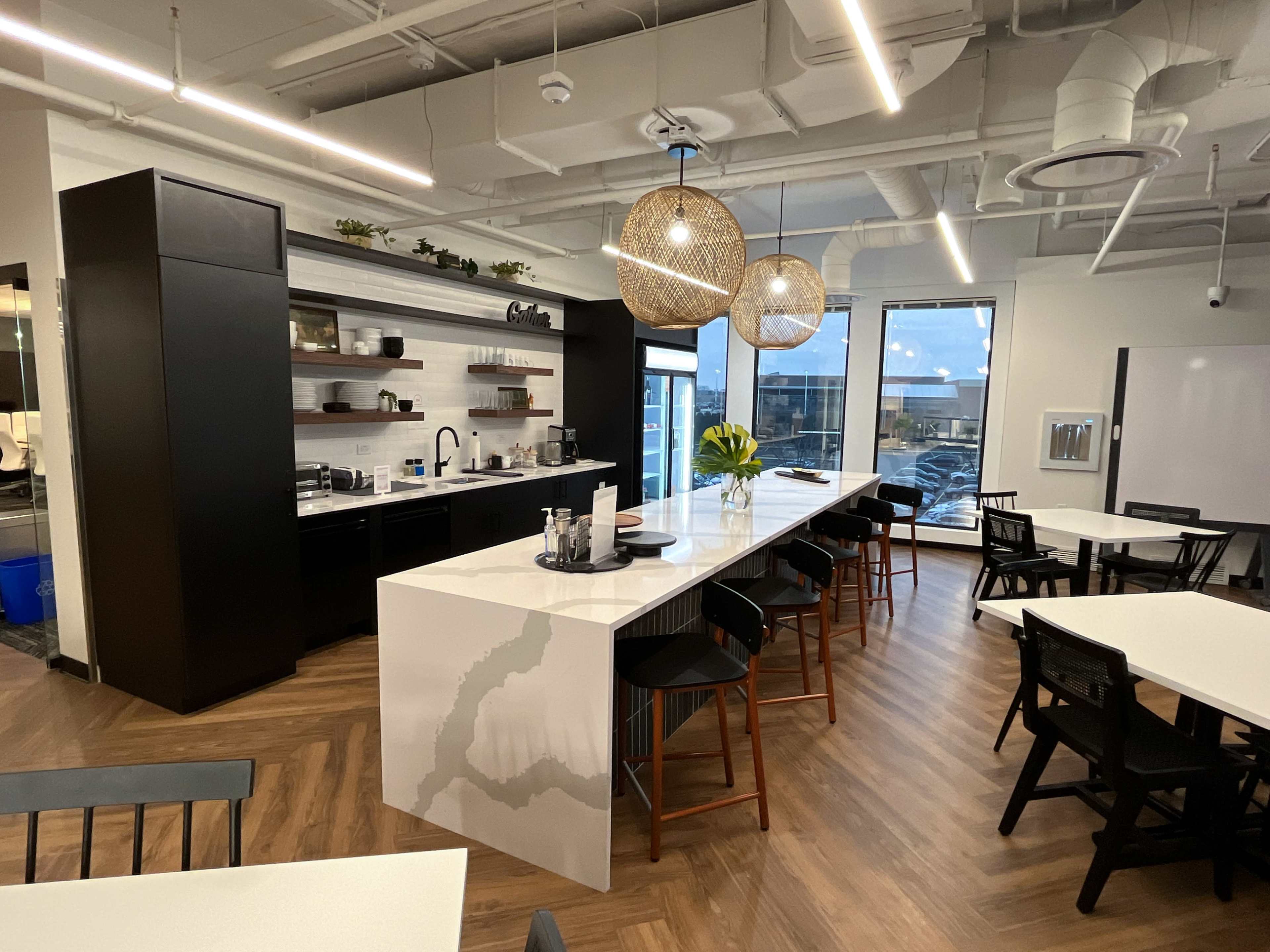 The image shows a modern kitchen area with a large marble countertop, black cabinetry, and pendant lighting, alongside multiple tables and chairs in an open space.
