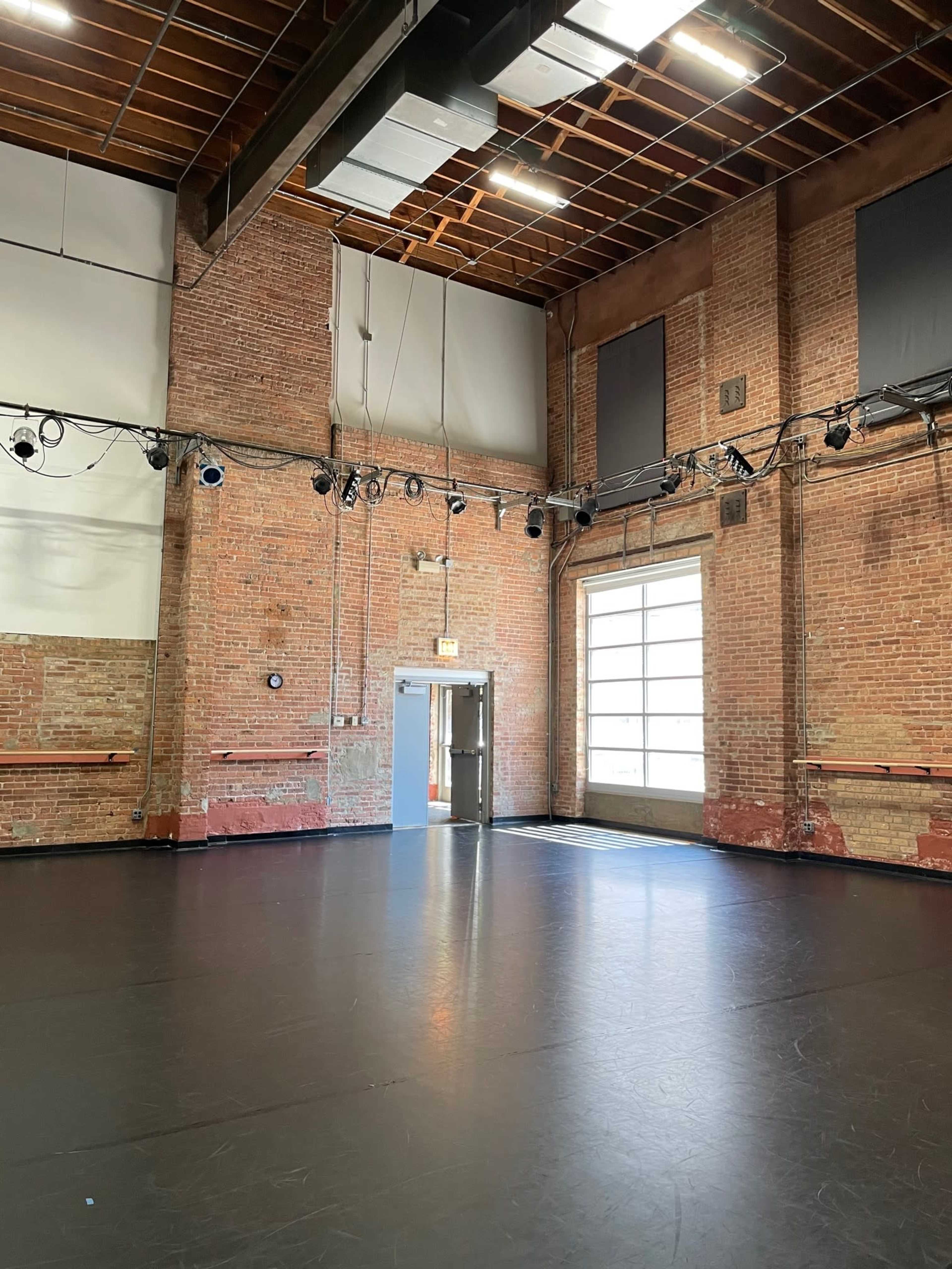 A spacious dance studio with exposed brick walls, wooden beams, and a large door leading outside.