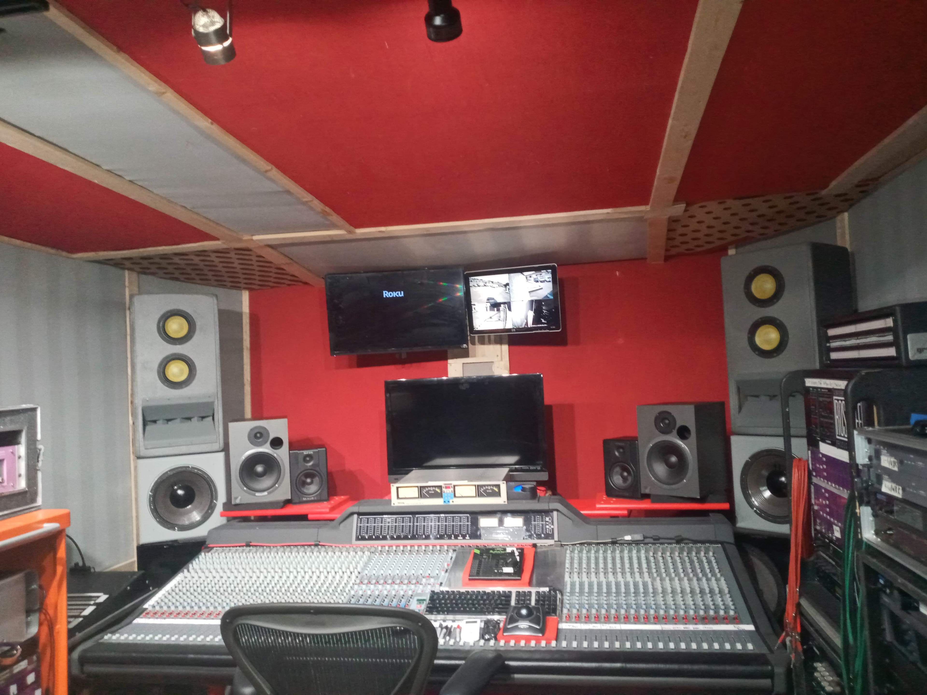 The image shows a sound recording studio featuring a large mixing console, multiple speakers, and two flat-screen monitors mounted on the walls.