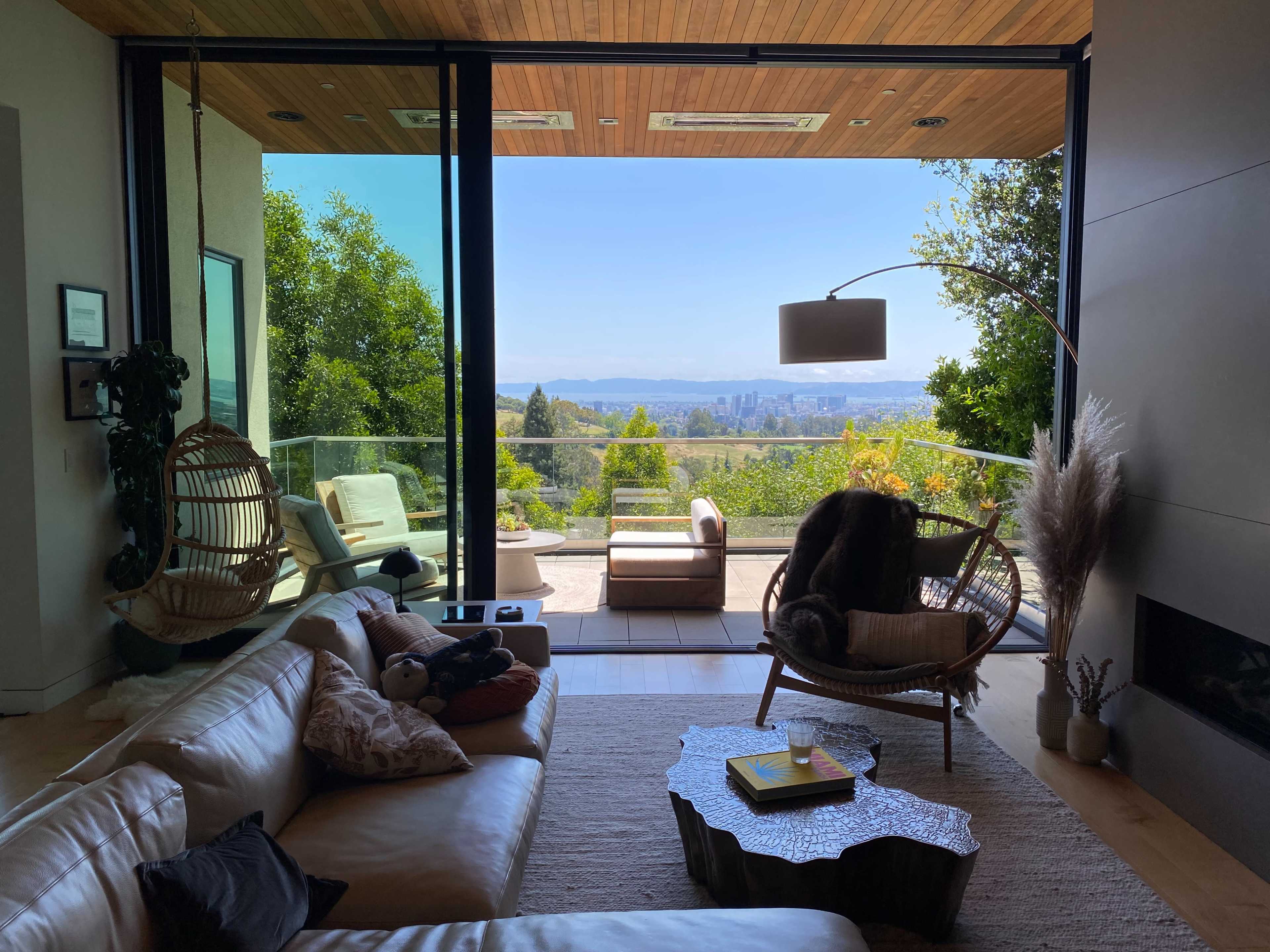 A modern living room features large windows that provide a view of a distant urban skyline, with a hanging chair and a plush sofa arranged around a decorative coffee table.