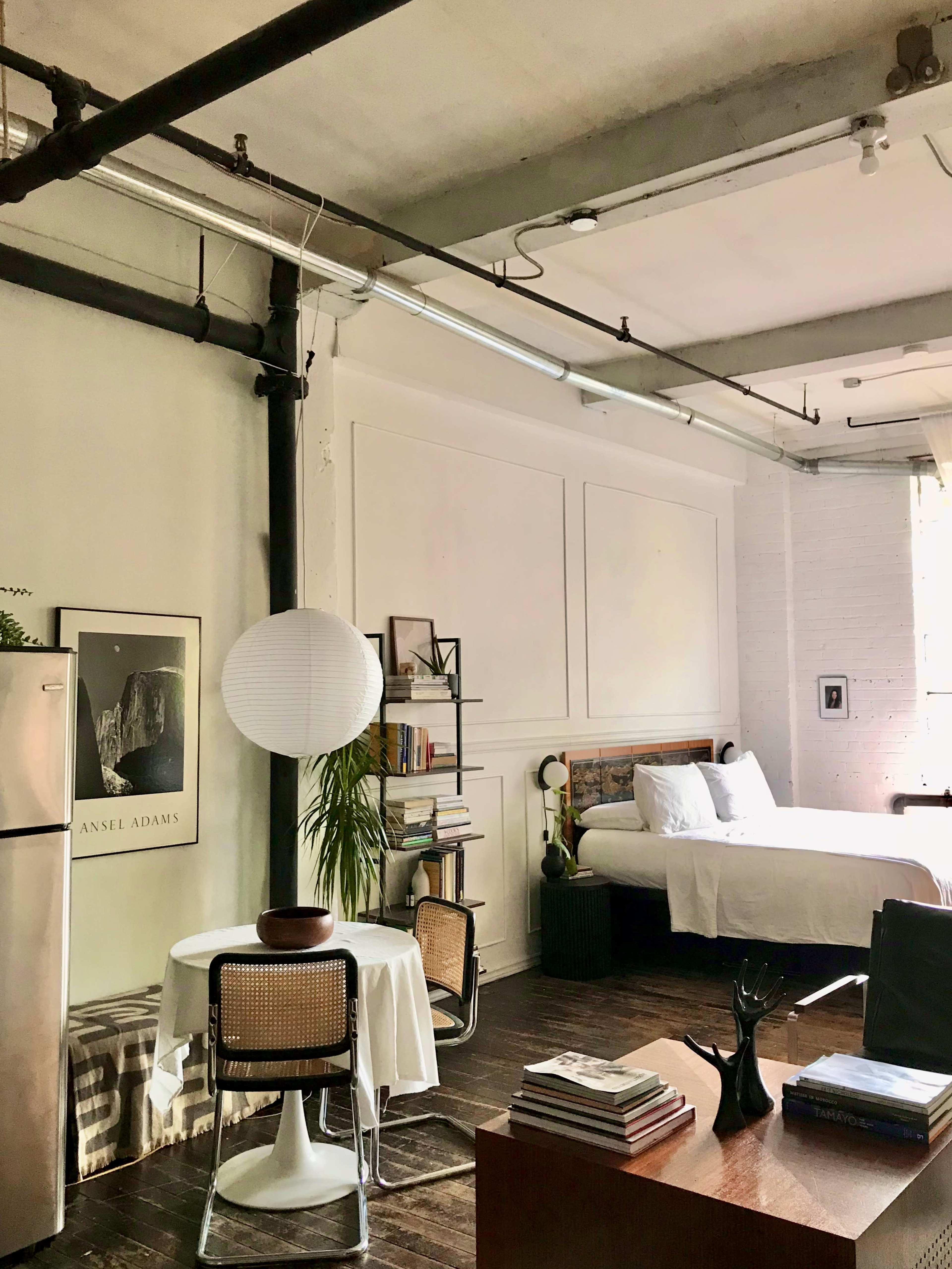 A modern studio apartment featuring wooden floors, a bed, a dining table with chairs, a small kitchenette, and decorative shelves.