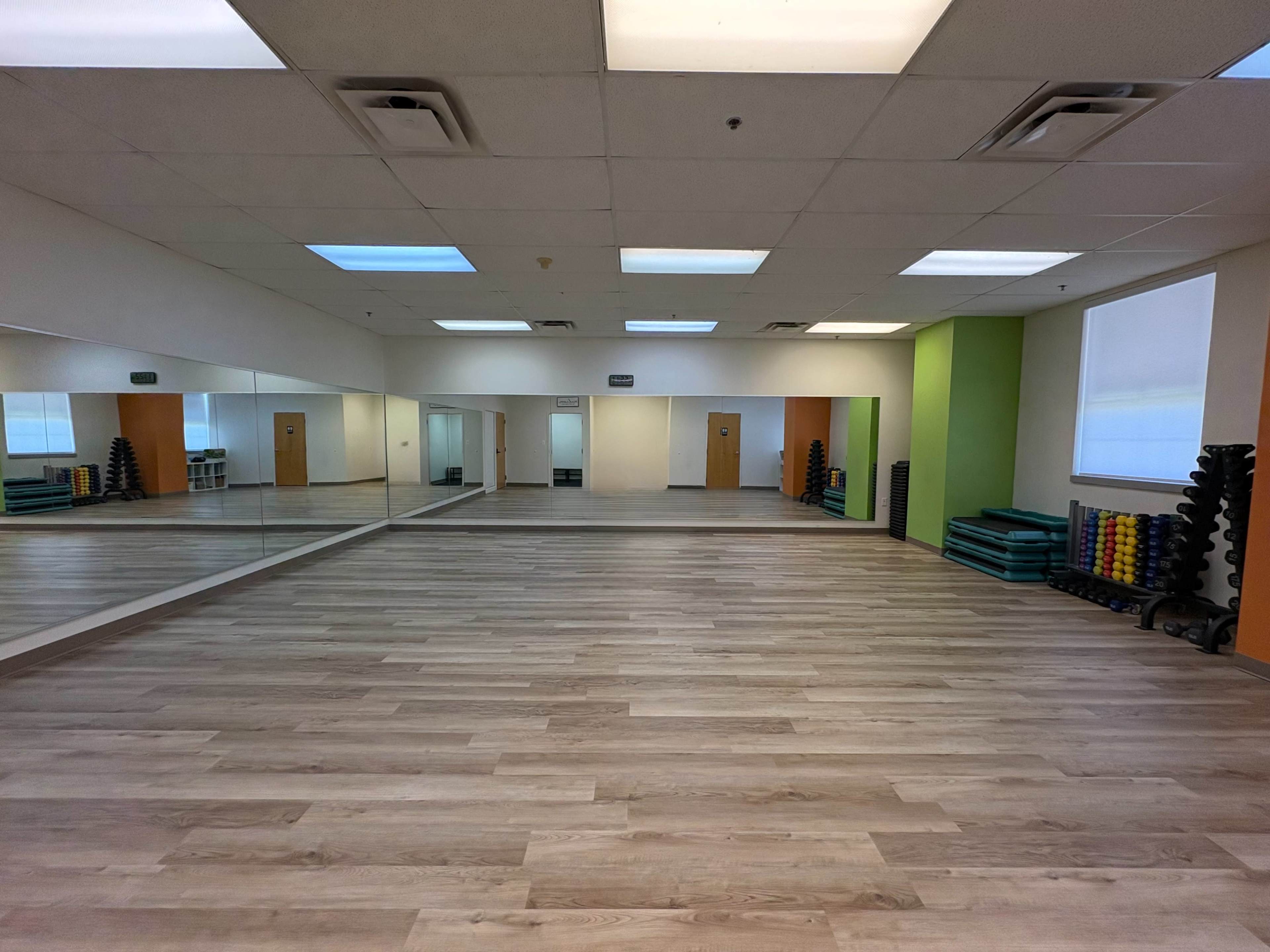 The image shows an empty fitness studio with wooden flooring, mirrors on one wall, and stacked exercise equipment in the corners.