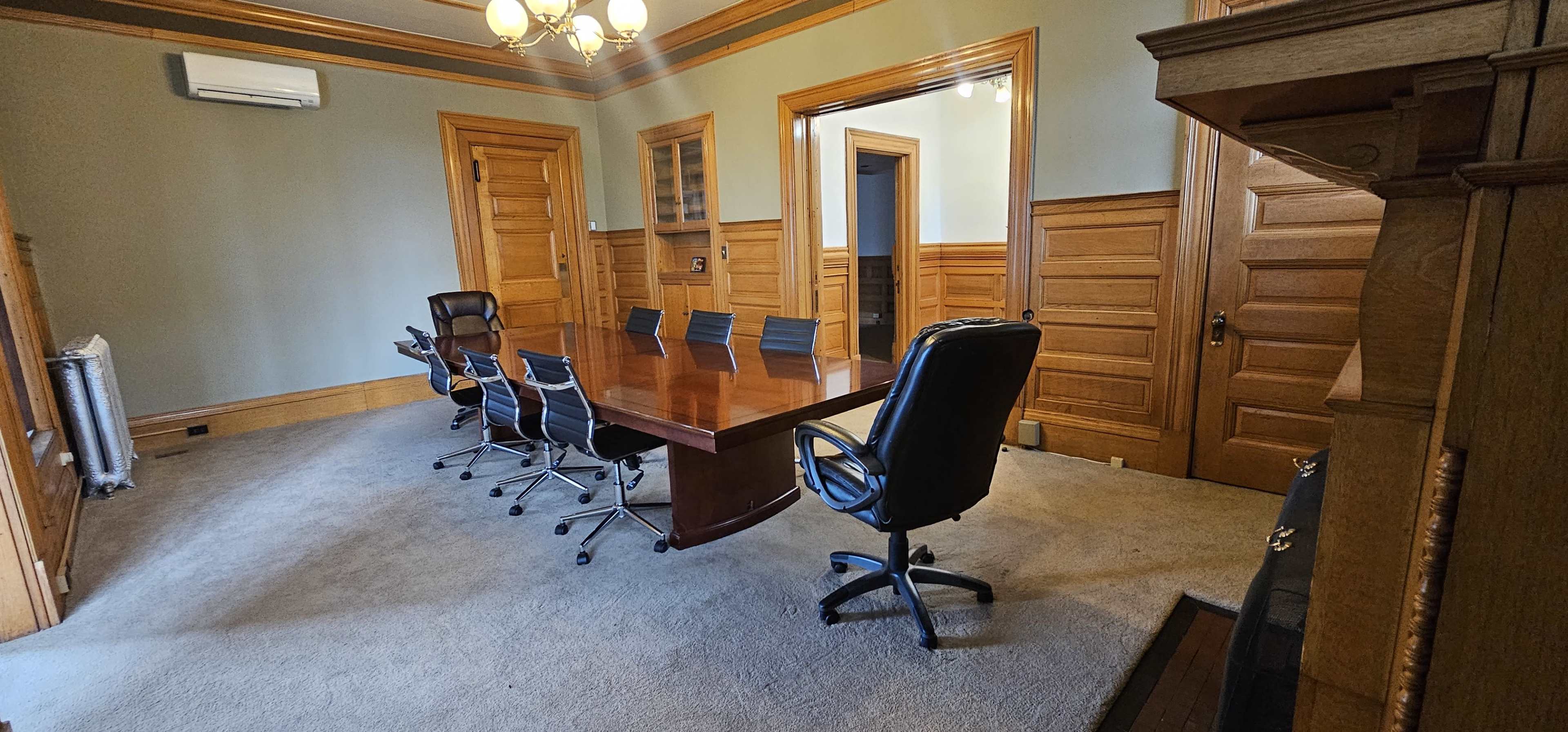 A spacious meeting room features a large wooden table surrounded by several office chairs, with wooden paneling and multiple doors leading to other areas.