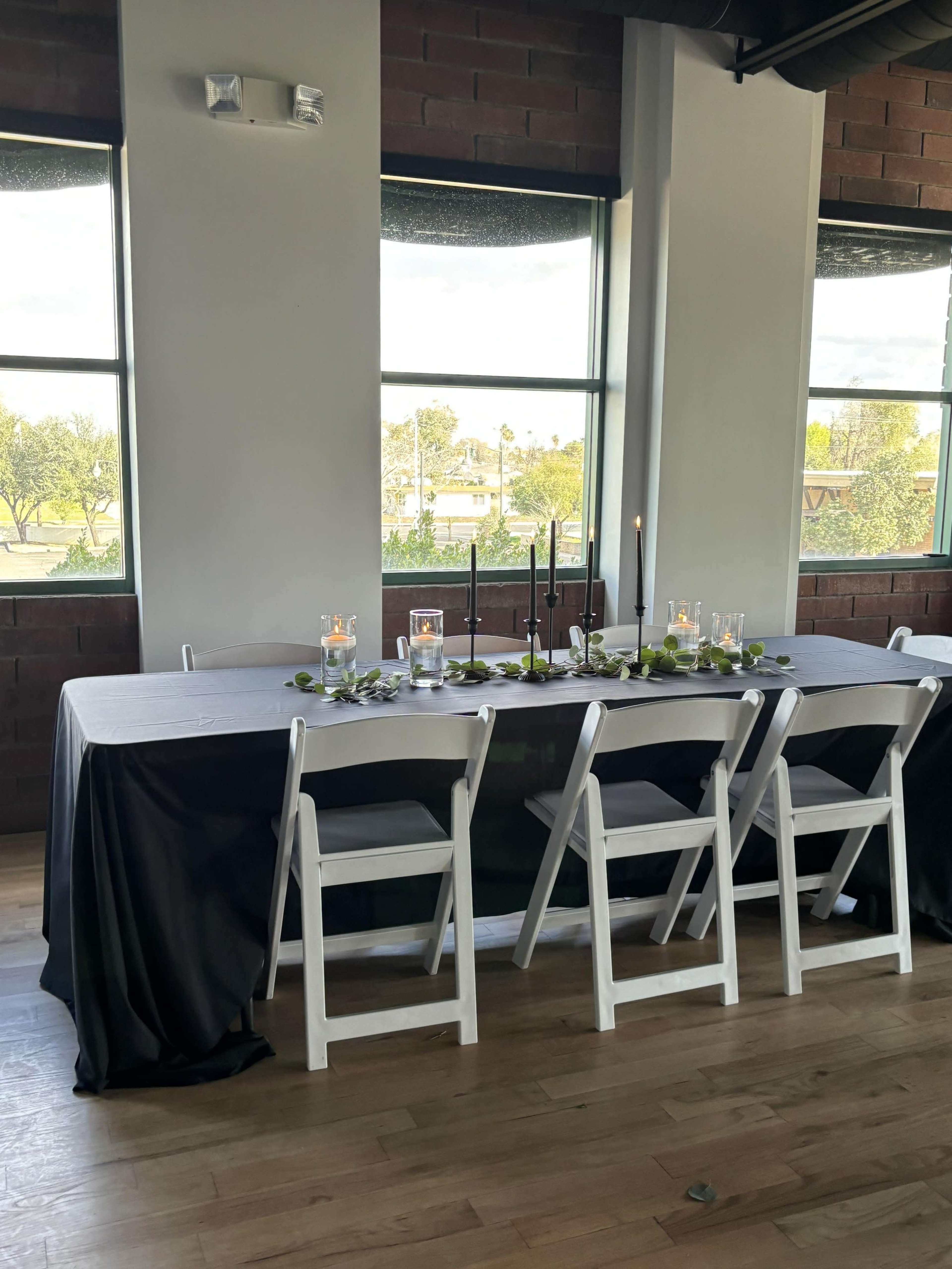 A long dining table is set up in a well-lit space with white and black tablecloths, surrounded by white chairs, and decorated with candles and greenery, with windows overlooking an outdoor area.