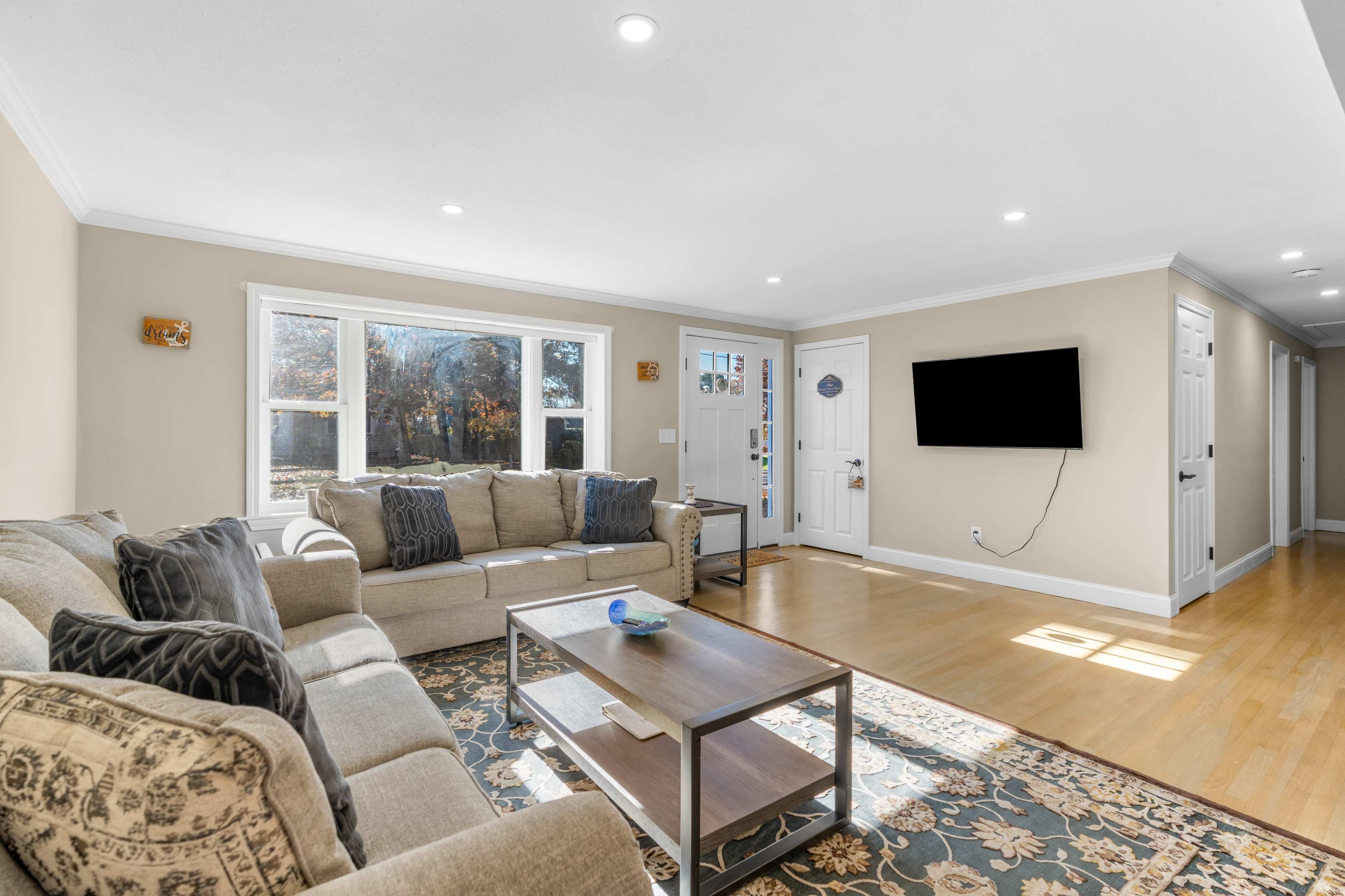 The living room features a beige sofa set arranged around a wooden coffee table, with large windows allowing natural light to fill the space.