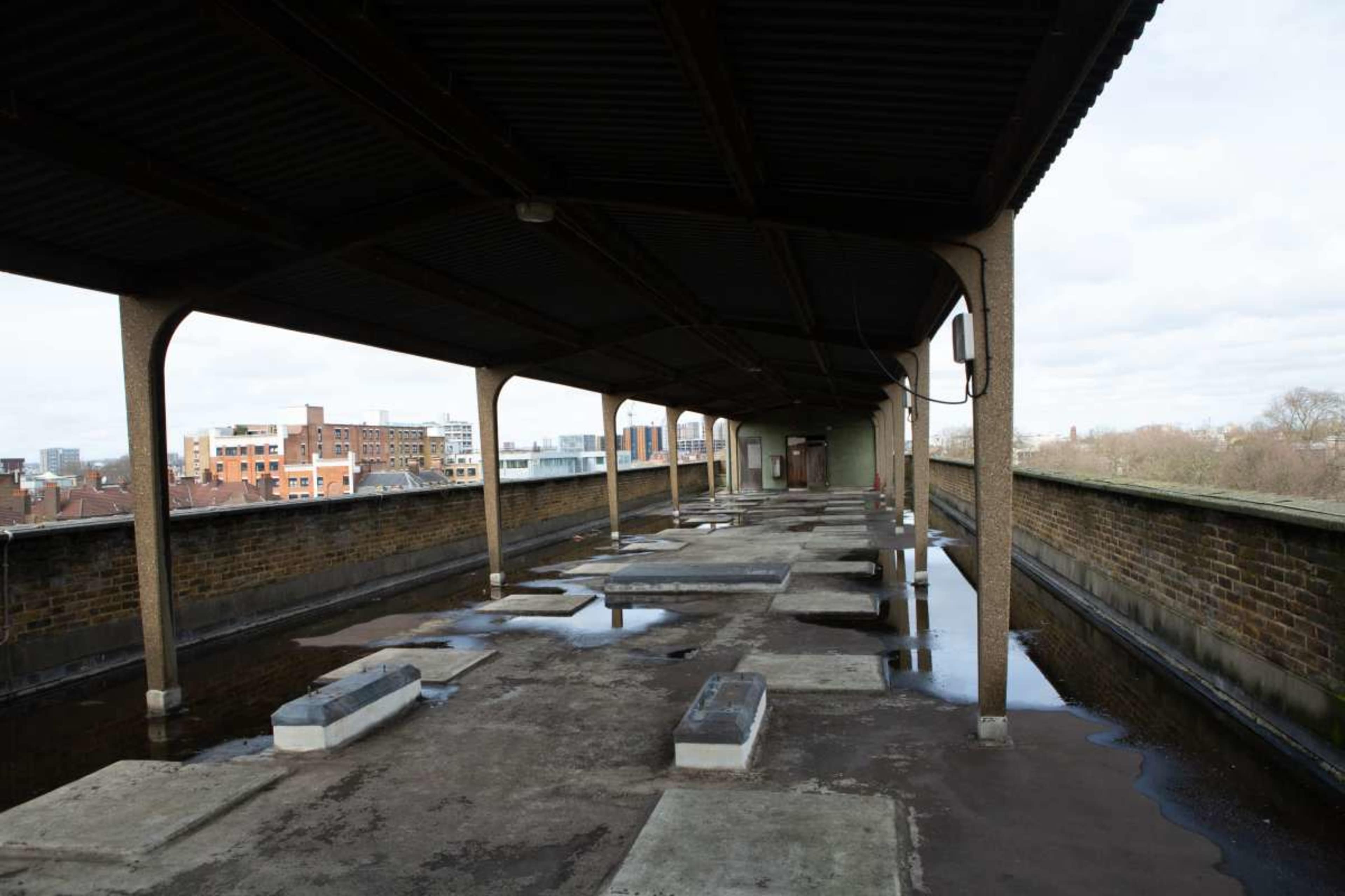 The image shows a vacant, covered walkway on a building's rooftop, featuring concrete slabs and puddles of water, with city buildings visible in the background.
