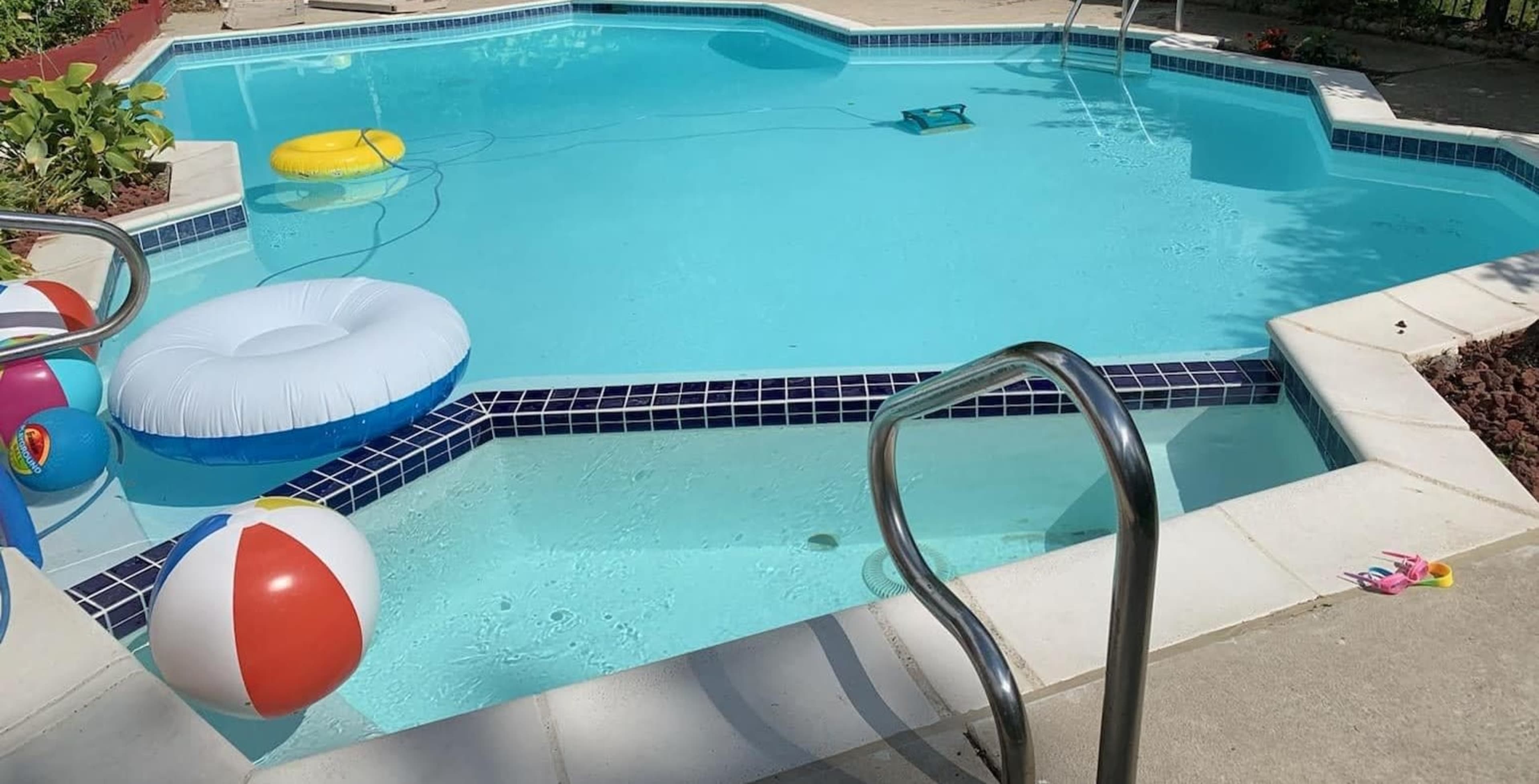 A swimming pool features colorful beach balls and inflatable floats, with a robotic cleaner in the water and steps leading into the pool.
