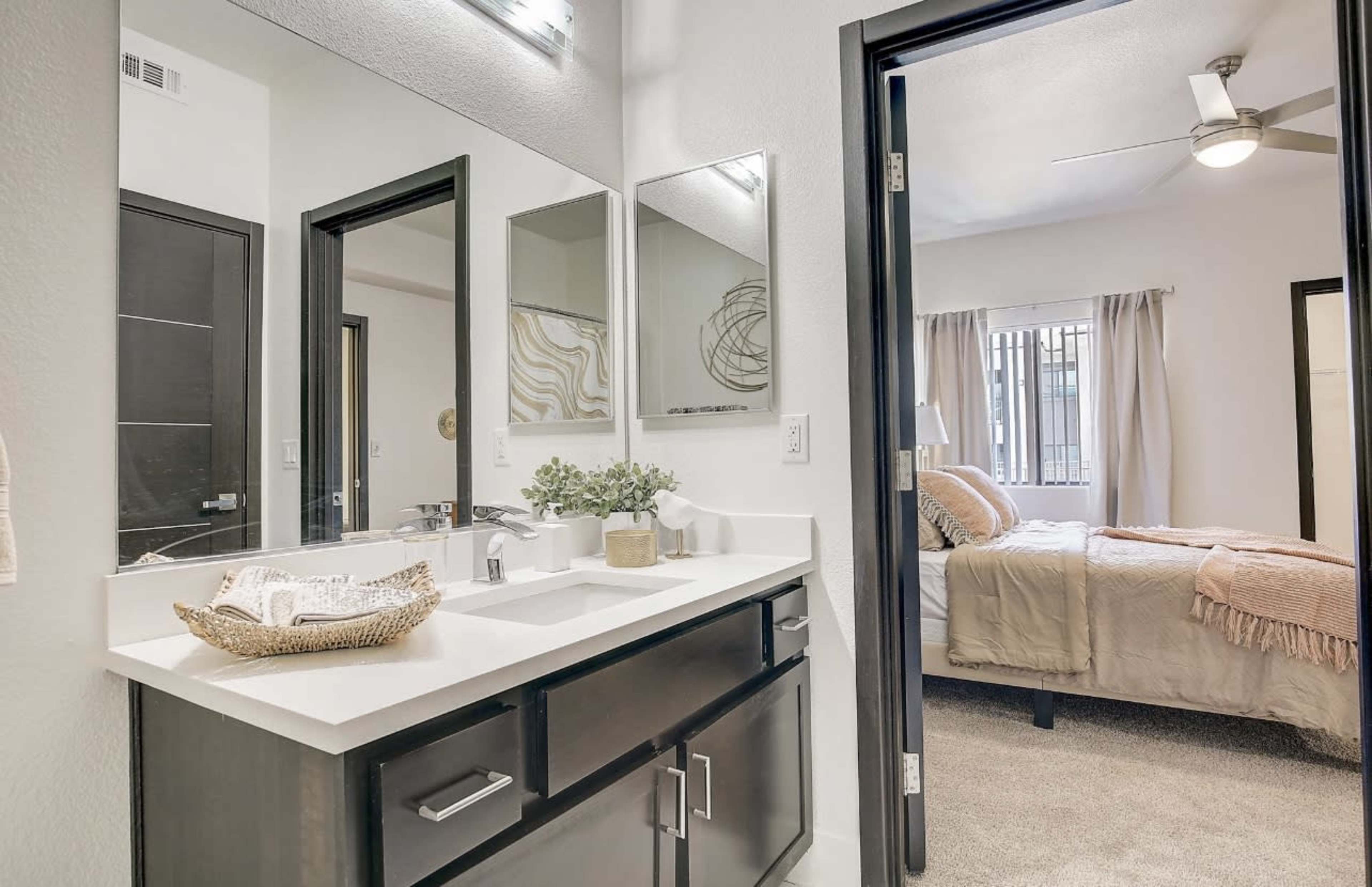 A bathroom with a double vanity and large mirrors, adjacent to a bedroom with a bed and window.