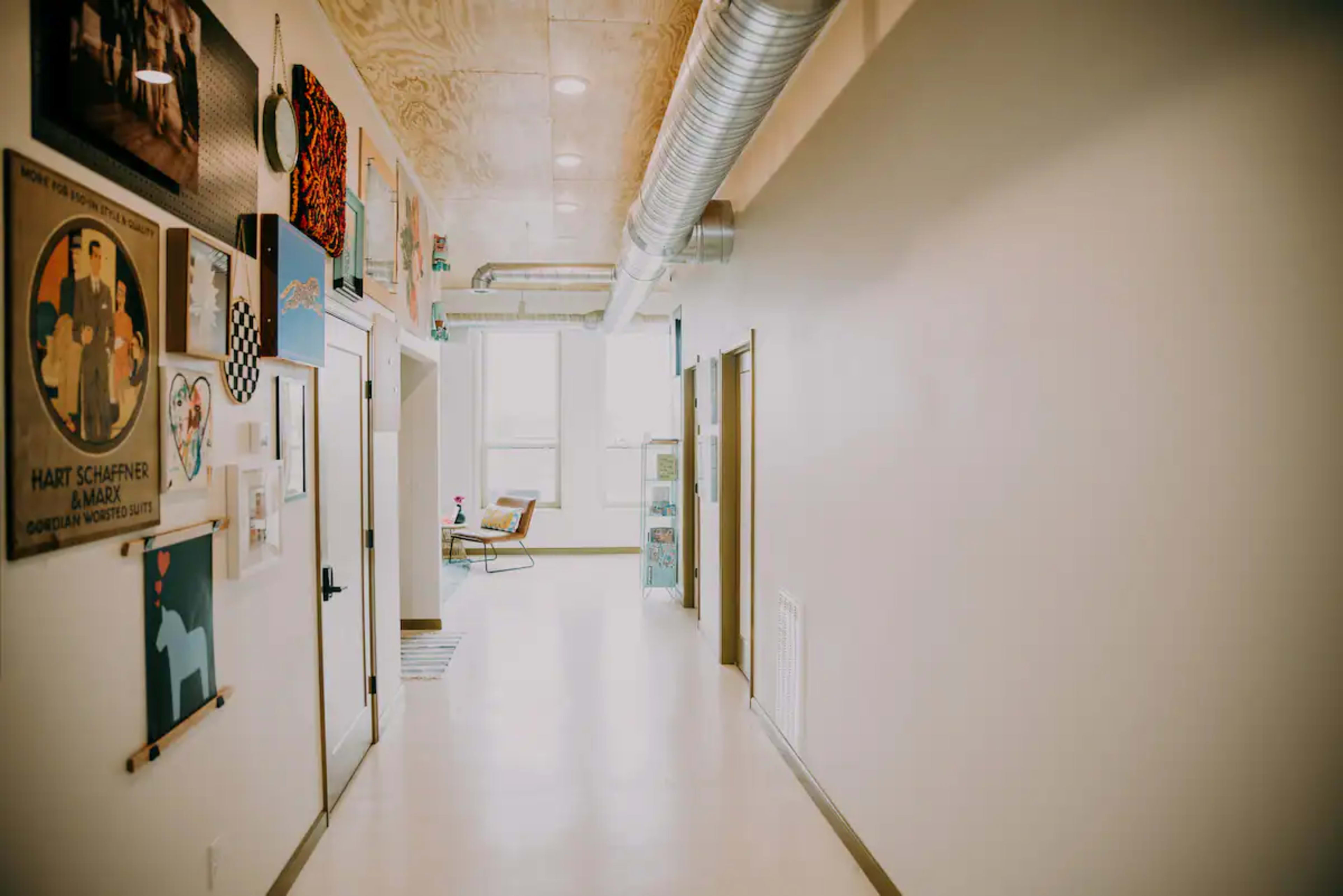 A long hallway features framed artwork on the walls, with doors leading to rooms and large windows at the far end.