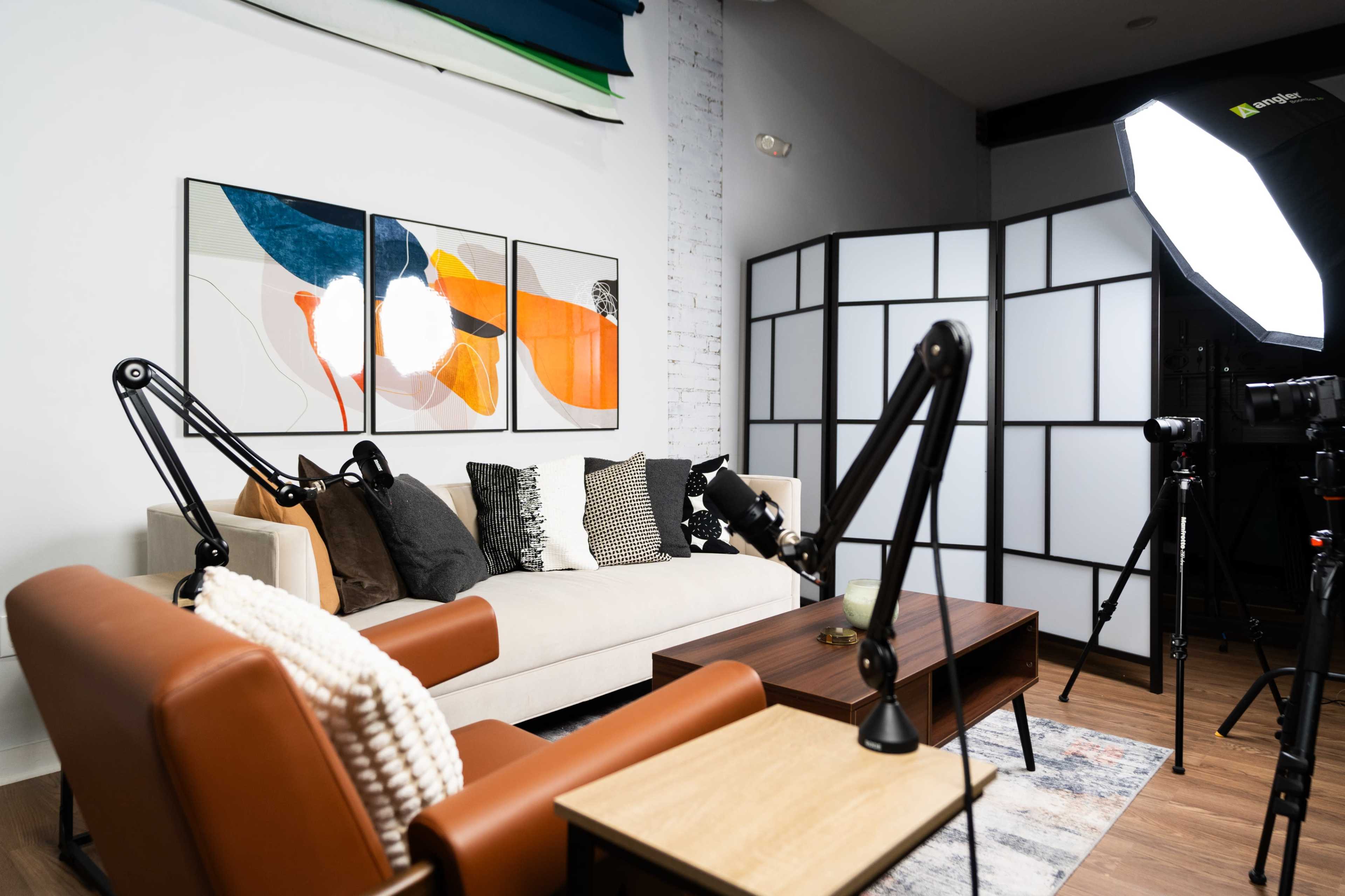 A modern living room setup with a beige sofa and armchairs, a wooden coffee table, and studio lighting equipment nearby.