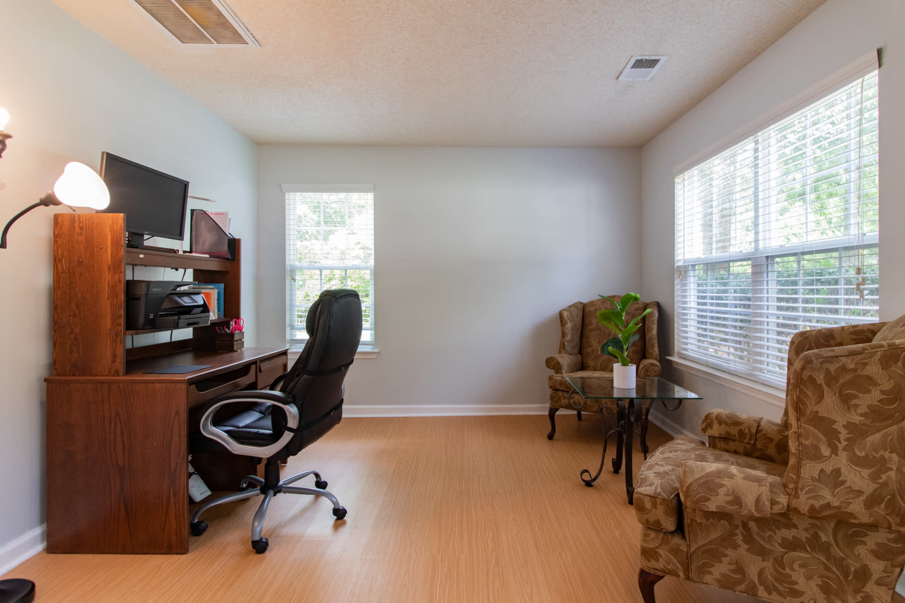 A home office with a wooden desk and chair, a computer, two upholstered armchairs, and a small table by a window.
