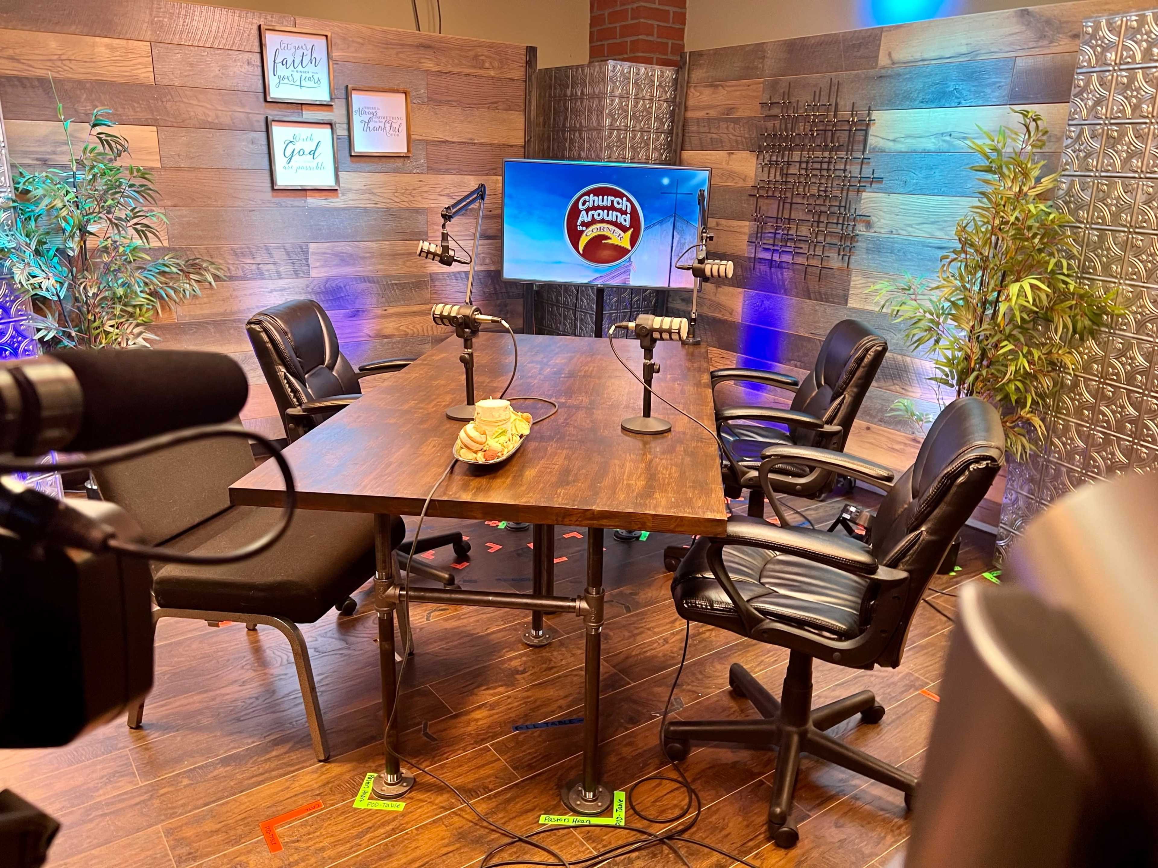 A media setup features a wooden table with four black chairs, microphones, and a screen displaying the "Church Growing" logo against a backdrop of wood paneling and decorative plants.