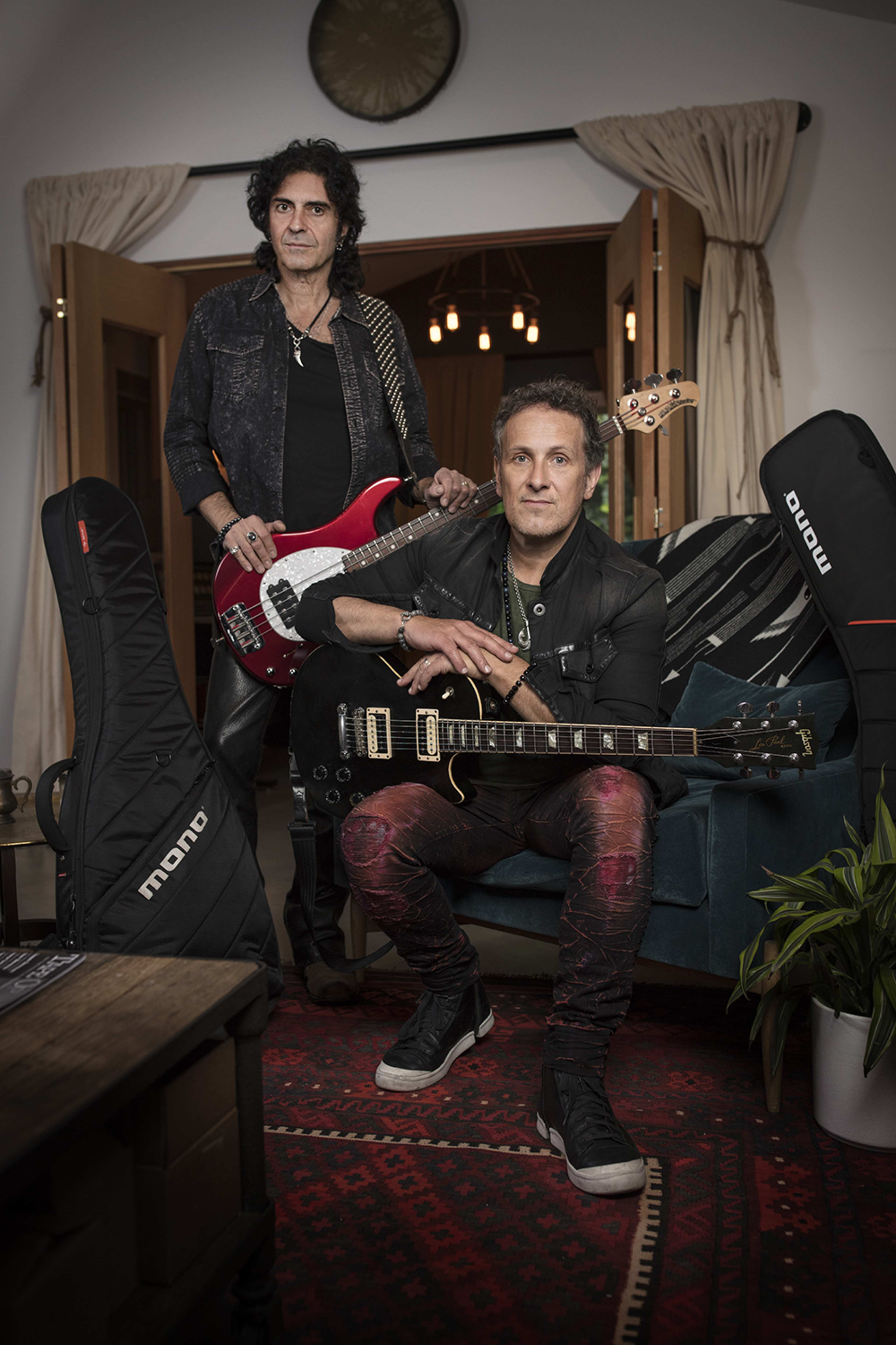 Two musicians pose in a room, one holding a red bass guitar and the other seated with a black electric guitar, surrounded by guitar cases and a cozy setting.