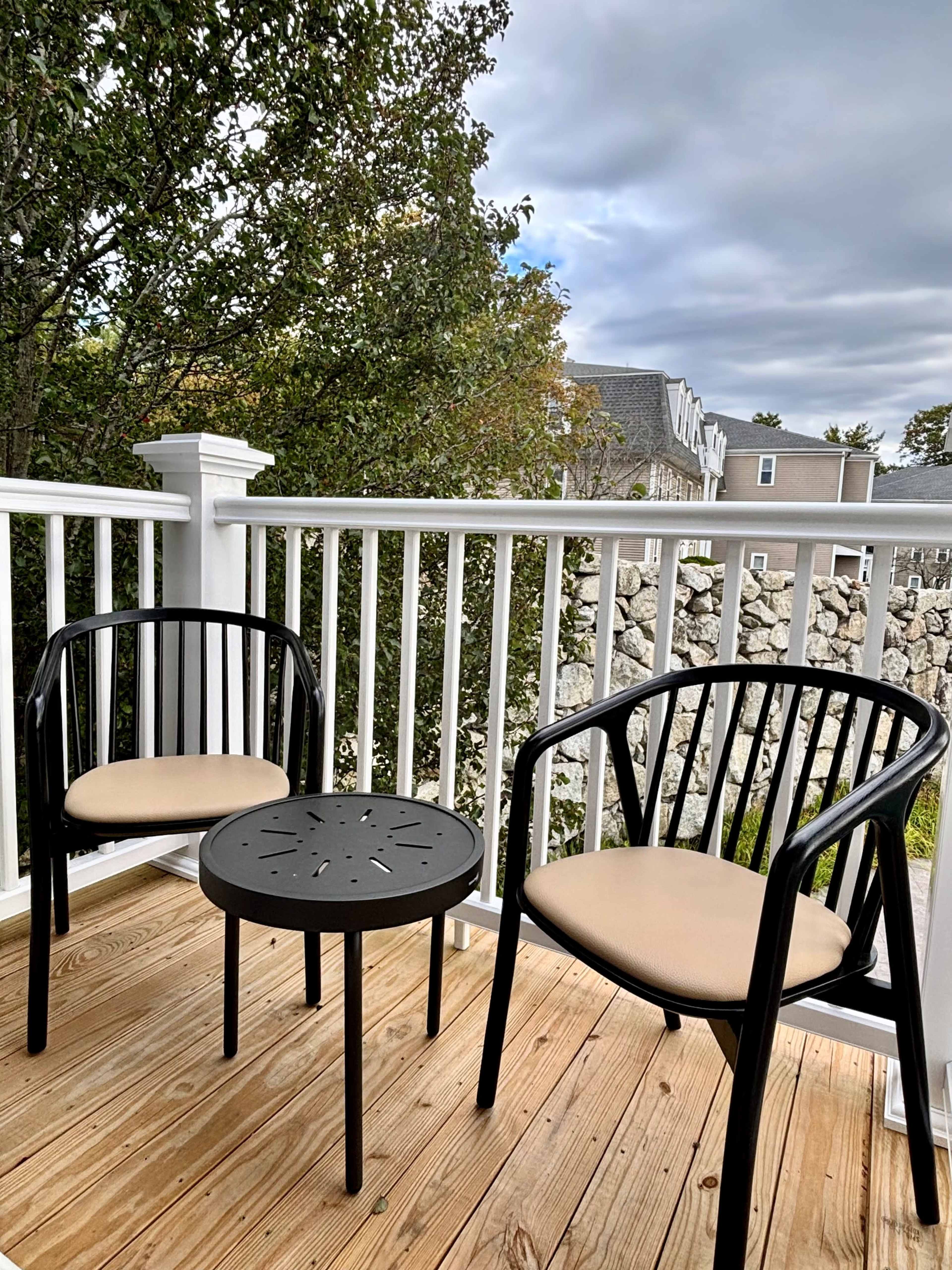 The image shows a small balcony with two black chairs and a round table on a wooden floor, surrounded by a white railing and greenery in the background.