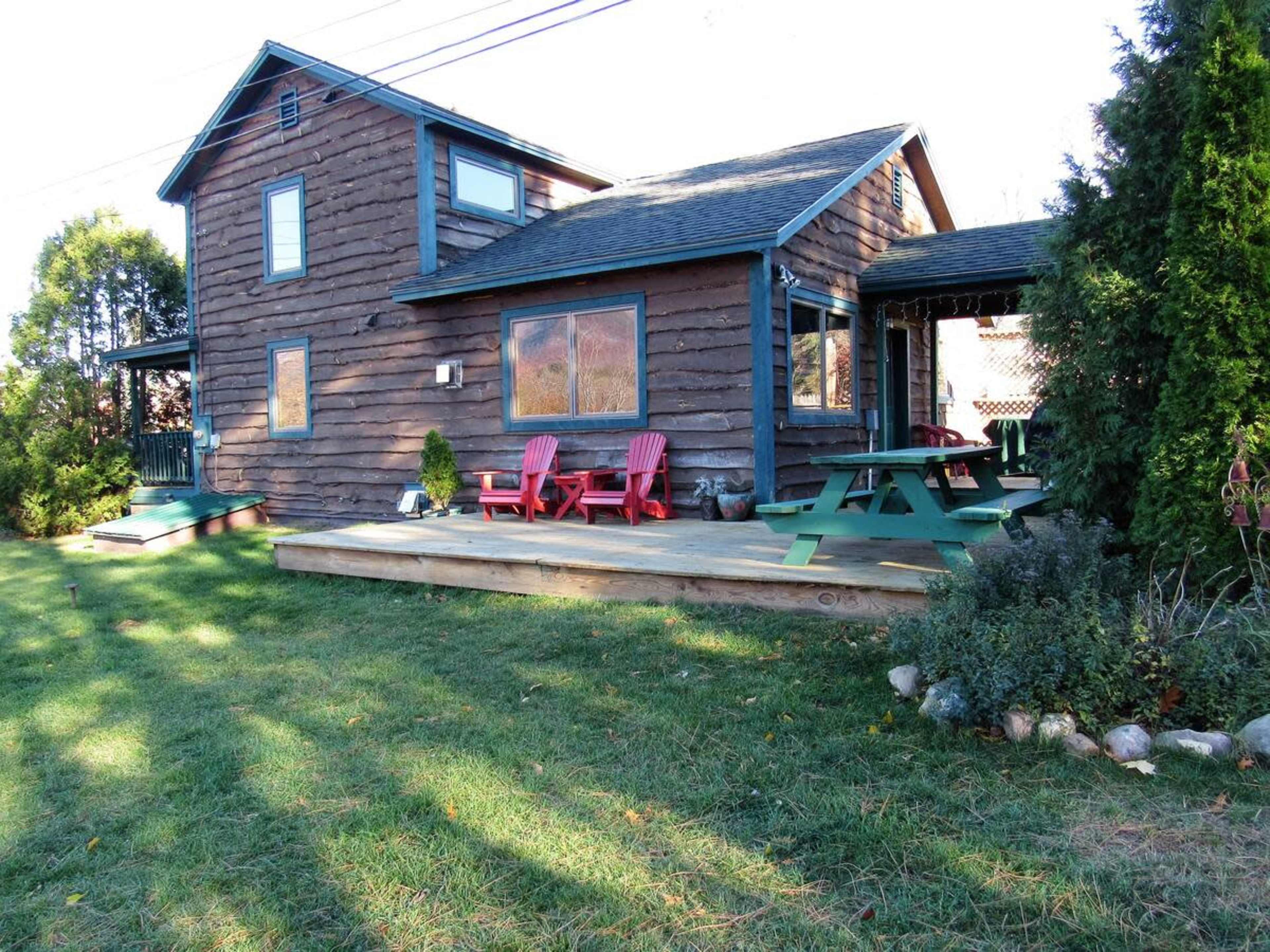 A log cabin with a wooden deck and red chairs sits surrounded by grass and trees.