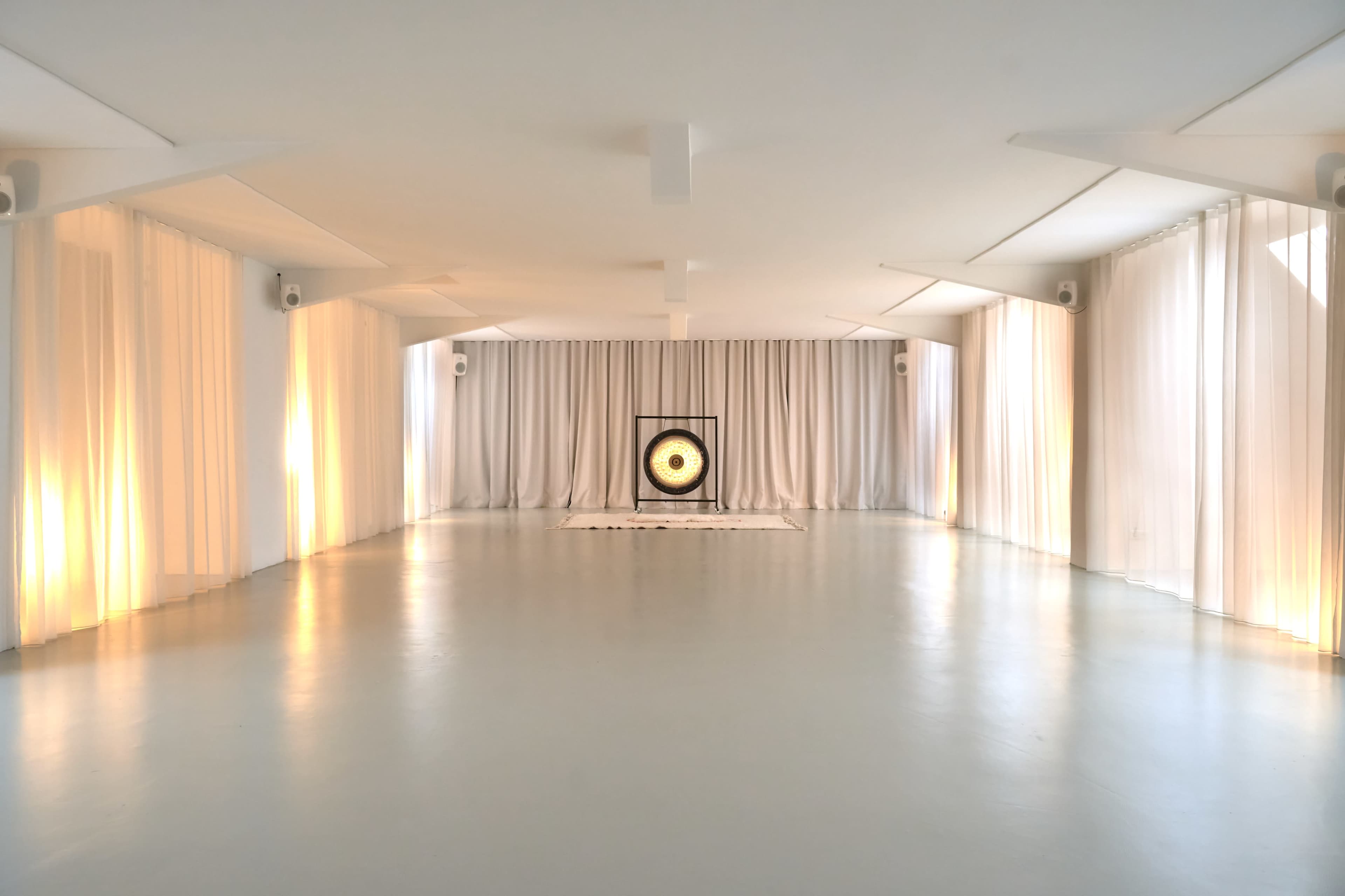 The image depicts a spacious, minimalist room with white curtains and a large gong centered against a backdrop of soft lighting.
