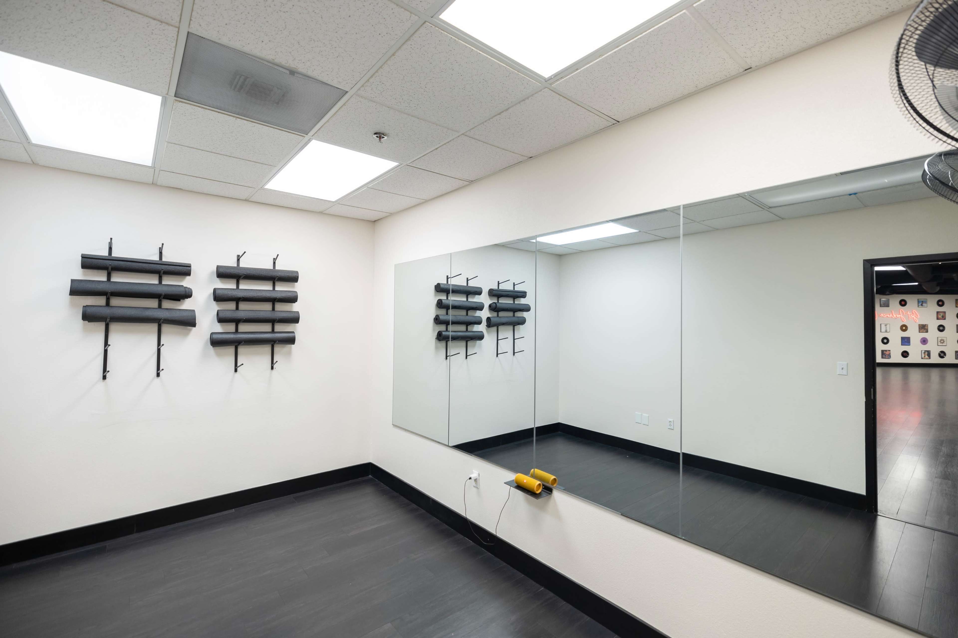 The image shows a sparsely decorated room with black wooden flooring, white walls, and a large mirror on one side, featuring wall-mounted black bars arranged in two clusters.