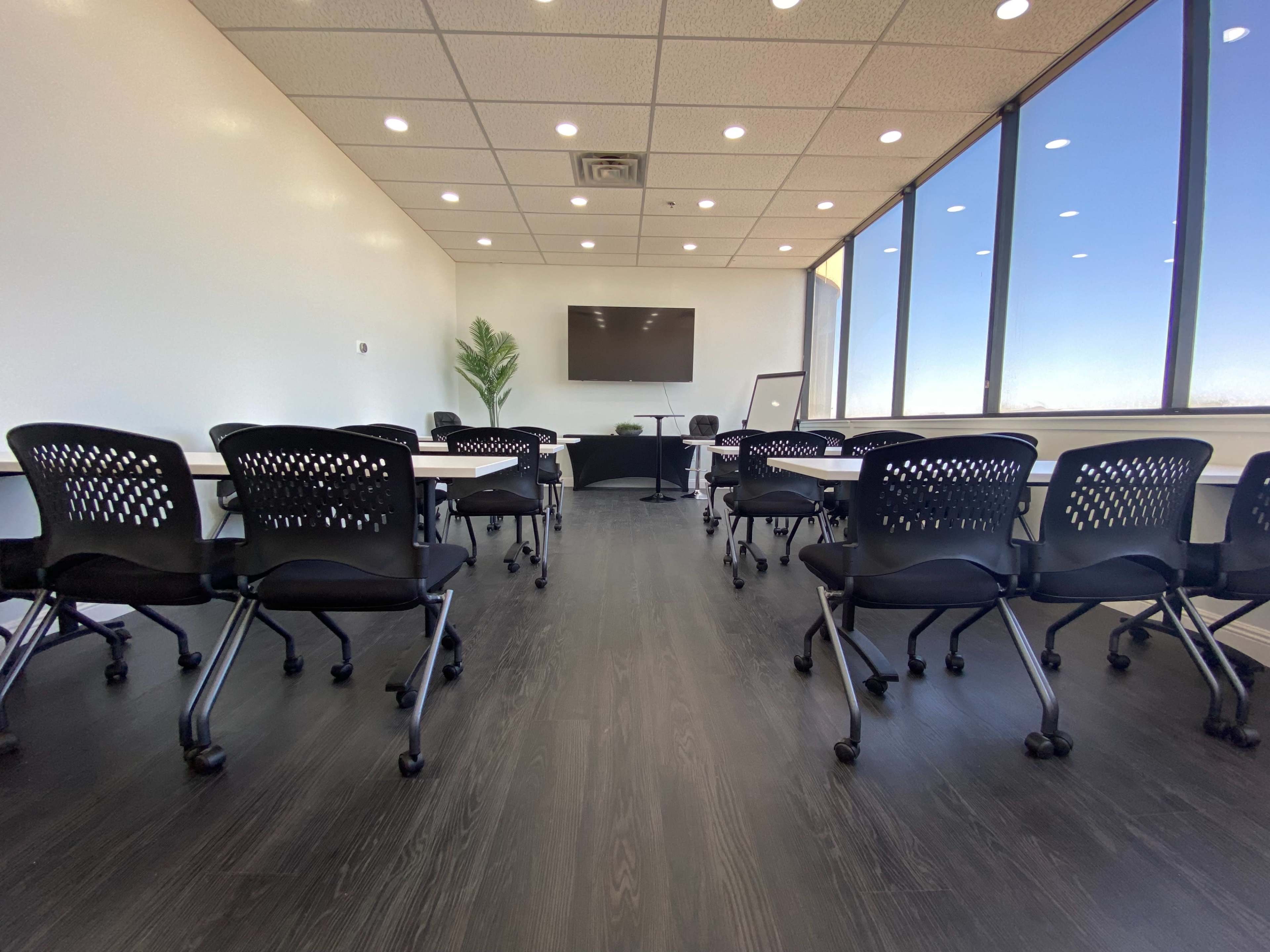 A modern conference room features several black rolling chairs arranged around tables, with large windows providing natural light and a view outside.