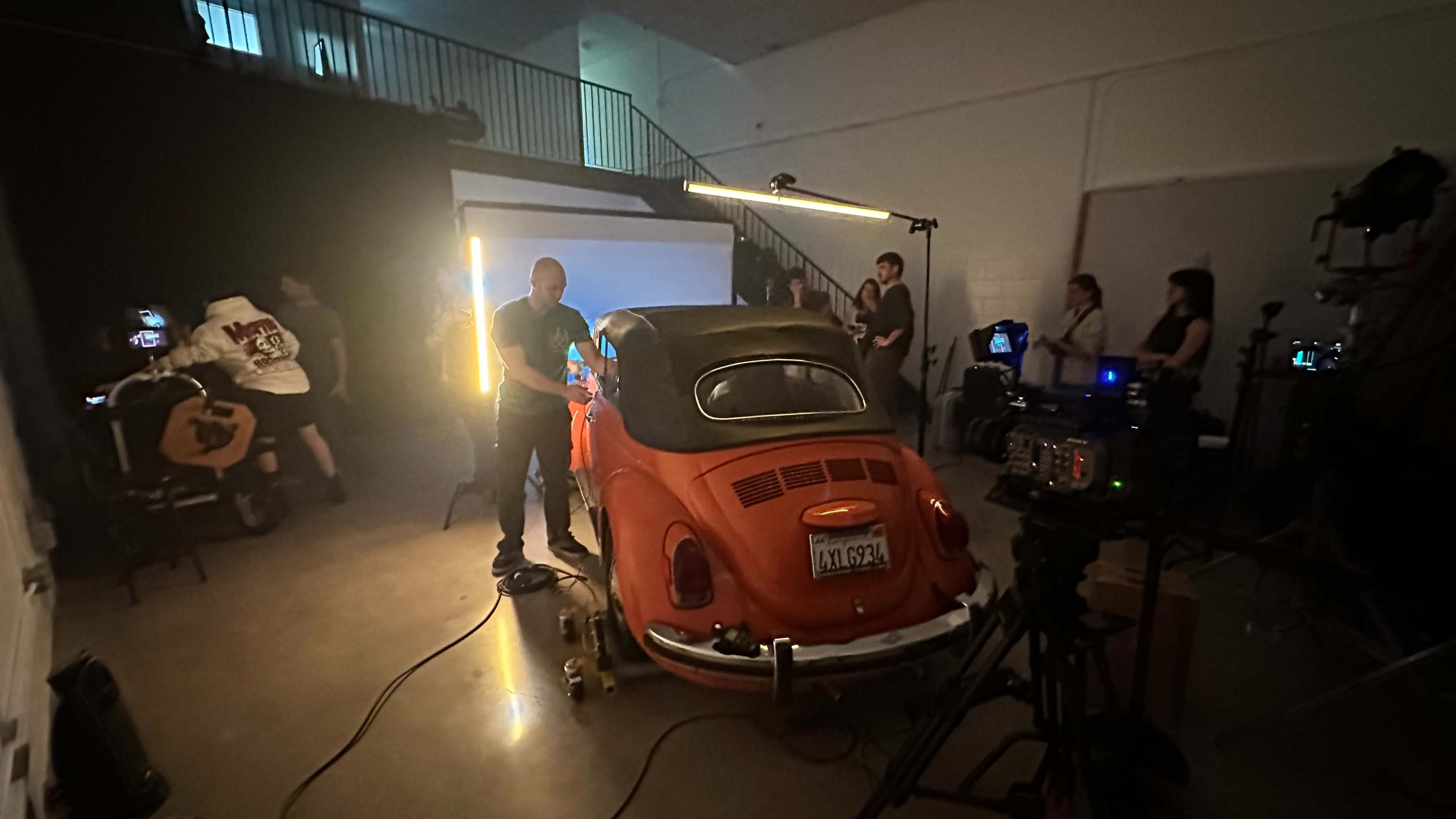 A group of people are working in a studio setting around an orange car on a wheeled platform, with cameras and lighting equipment set up for a shoot.