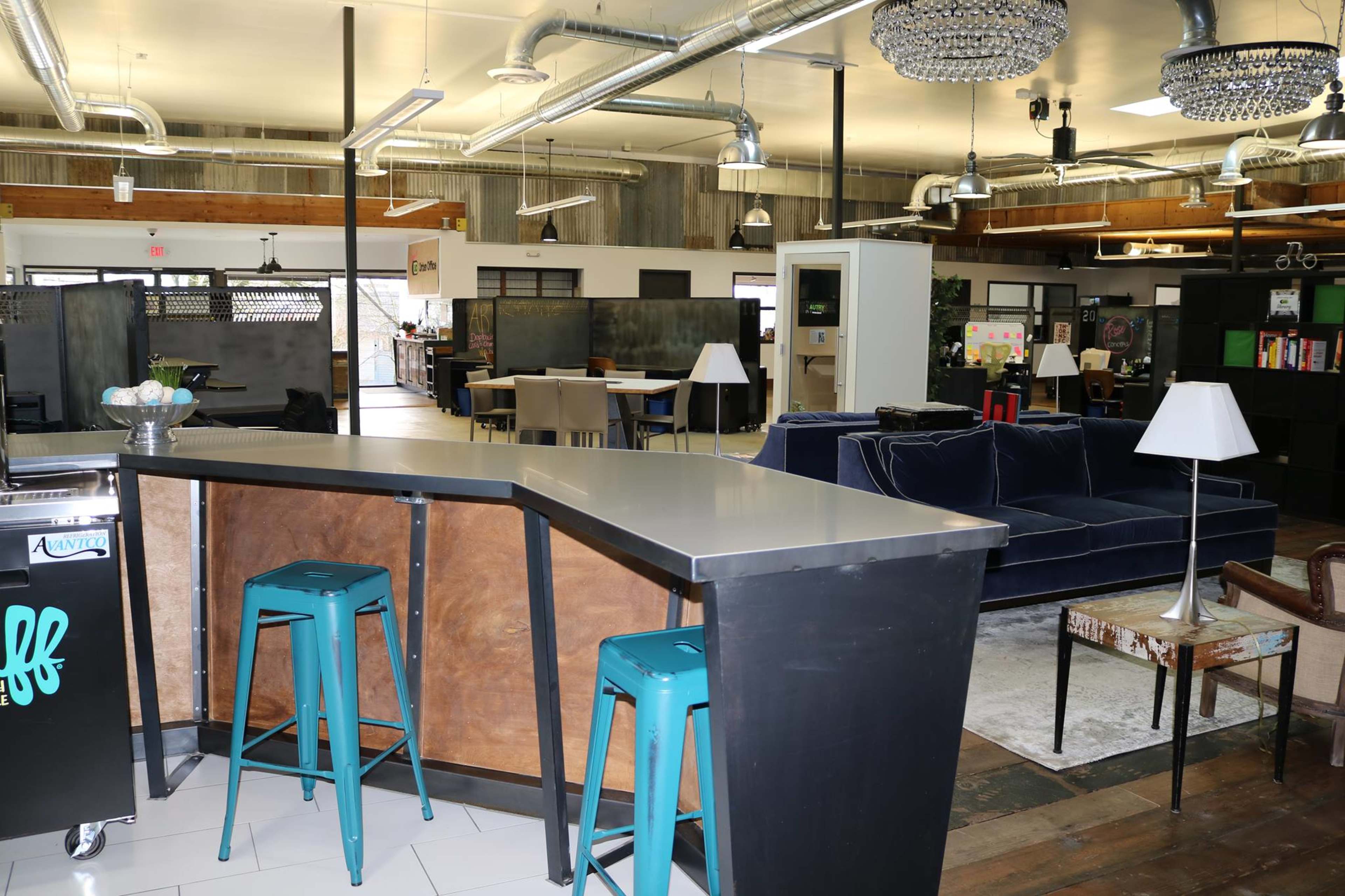 A spacious and modern workspace with a bar-style table, teal stools, and various seating areas in a well-lit environment.