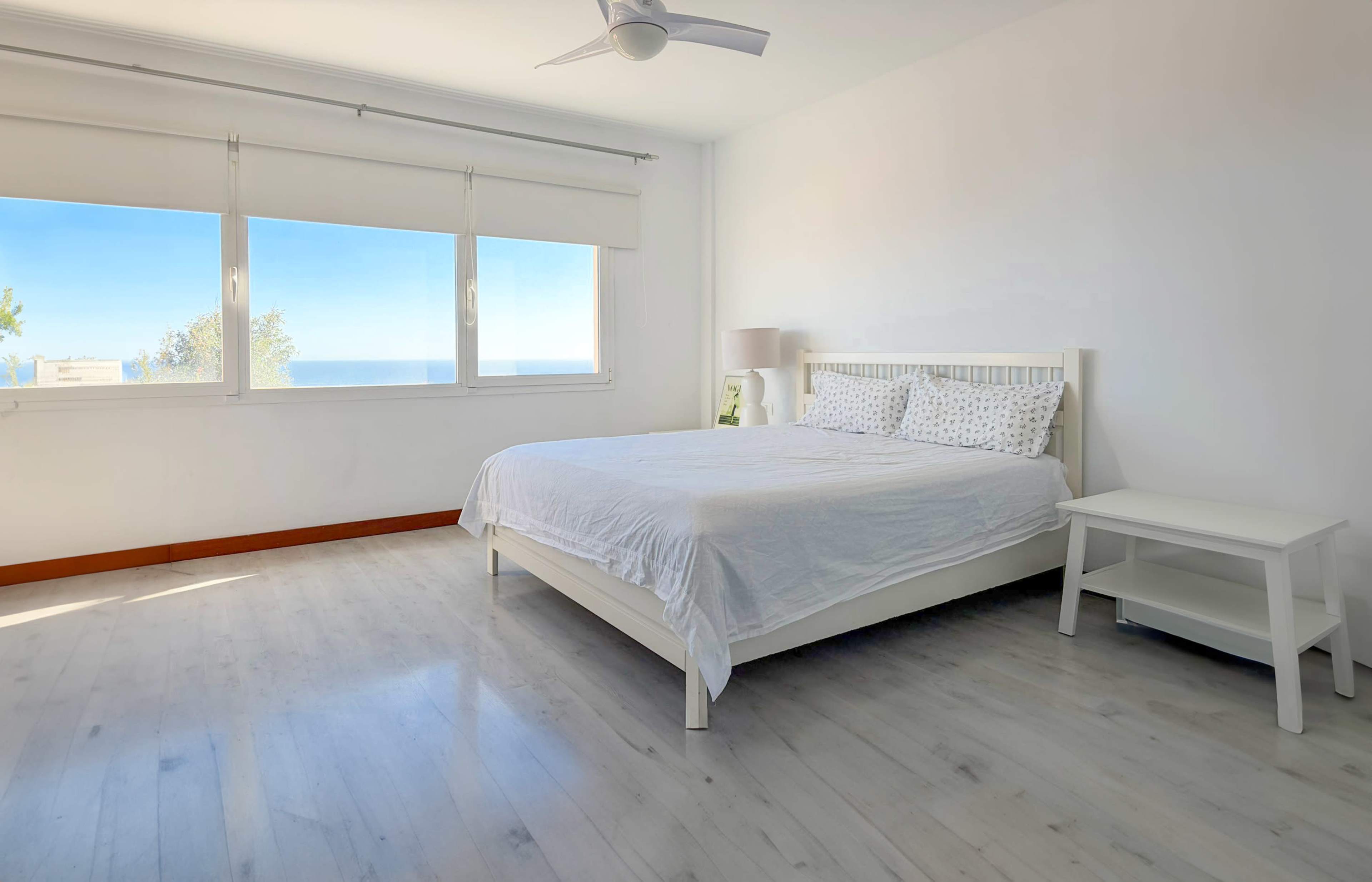 The image shows a bright, minimalist bedroom featuring a white bed, a bedside table, and large windows with an ocean view.