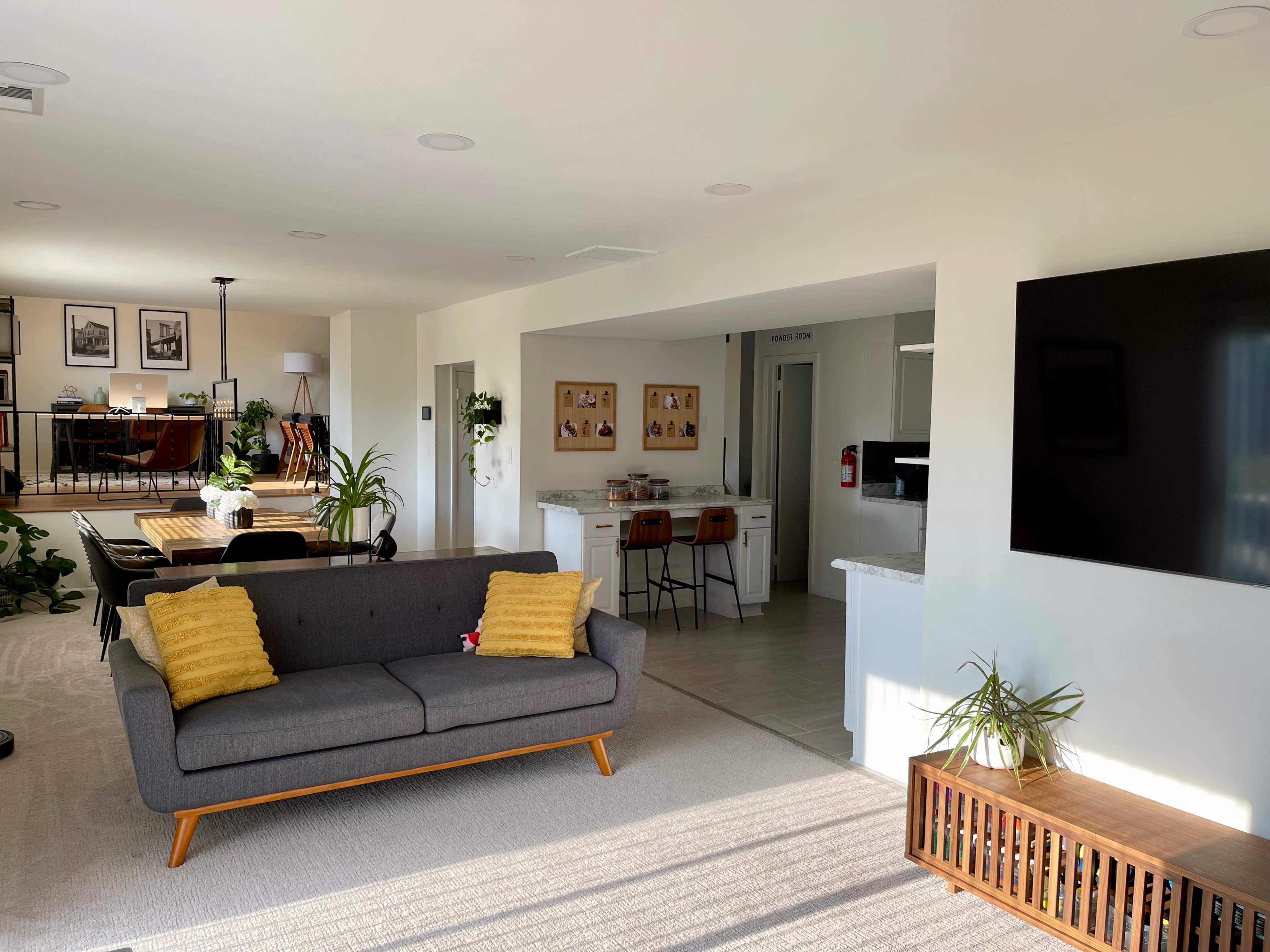 A modern living room featuring a gray sofa with yellow pillows, a wooden coffee table, and an open layout leading to a dining area and kitchen in the background.