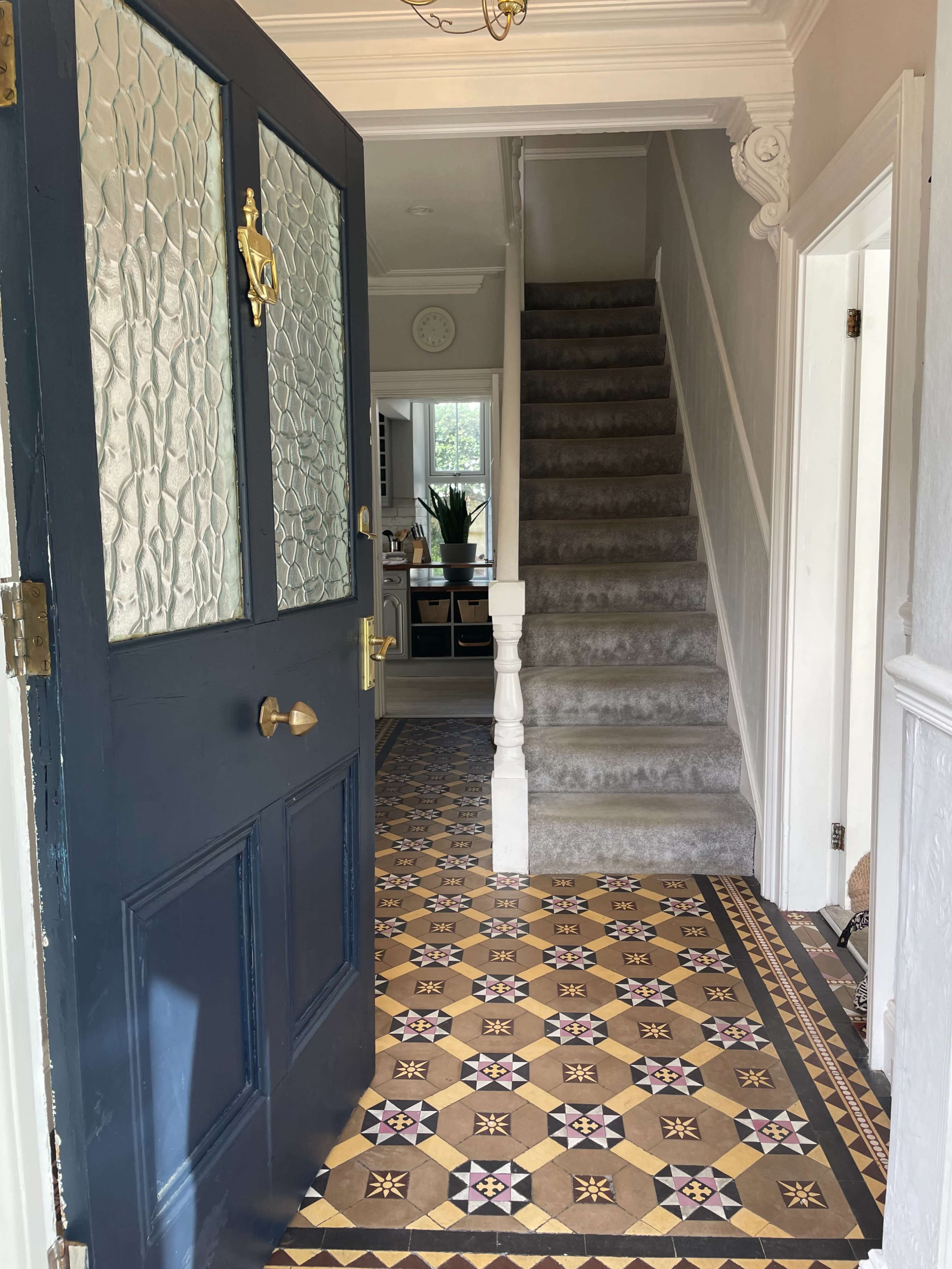 An open front door leading into a hallway with patterned tile flooring and a staircase in the background.