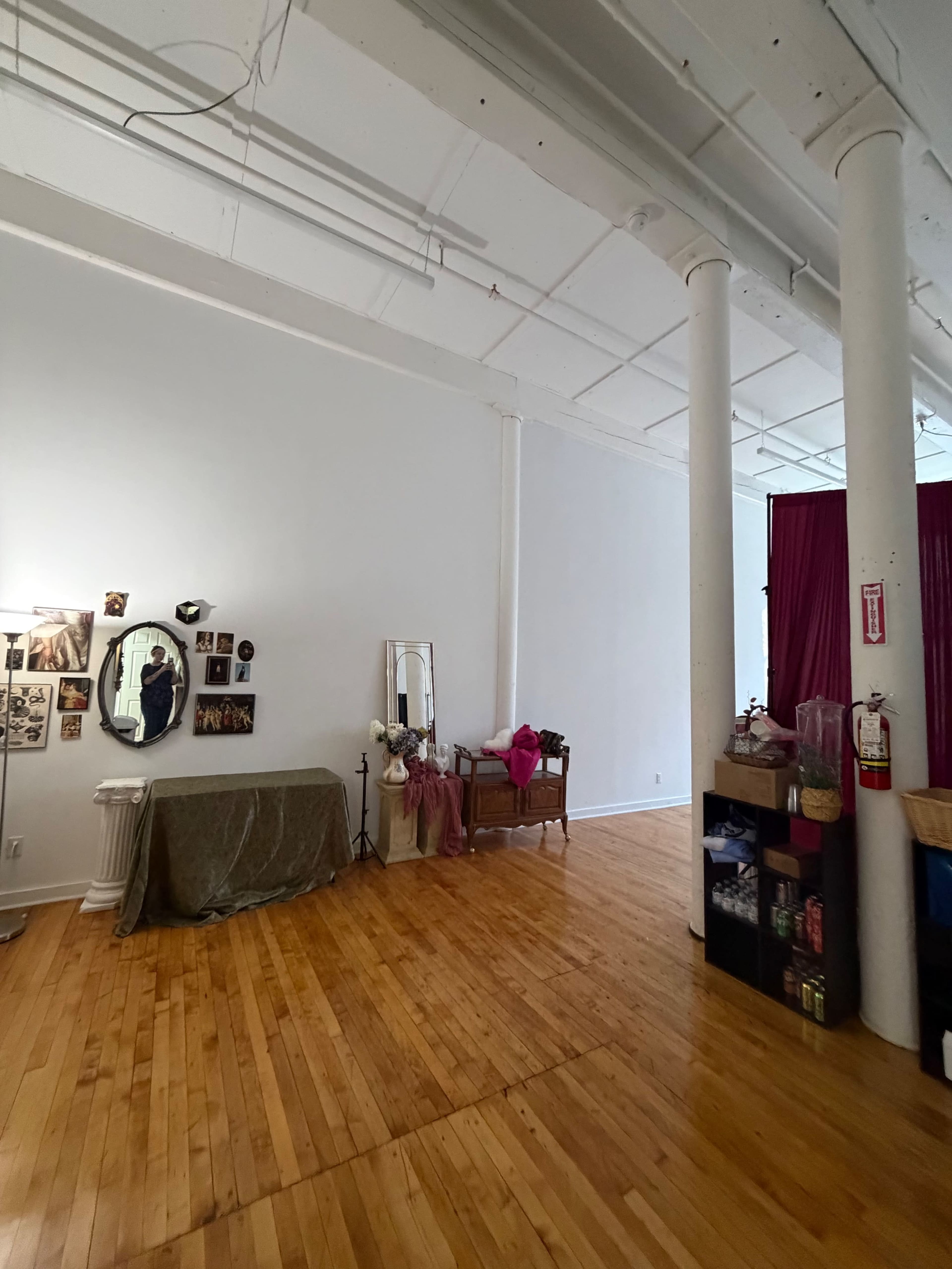 The image shows a spacious, empty room with wooden floors, white walls, and two large columns, featuring a mirror and photographs on one wall and a table with decorative items on another side.
