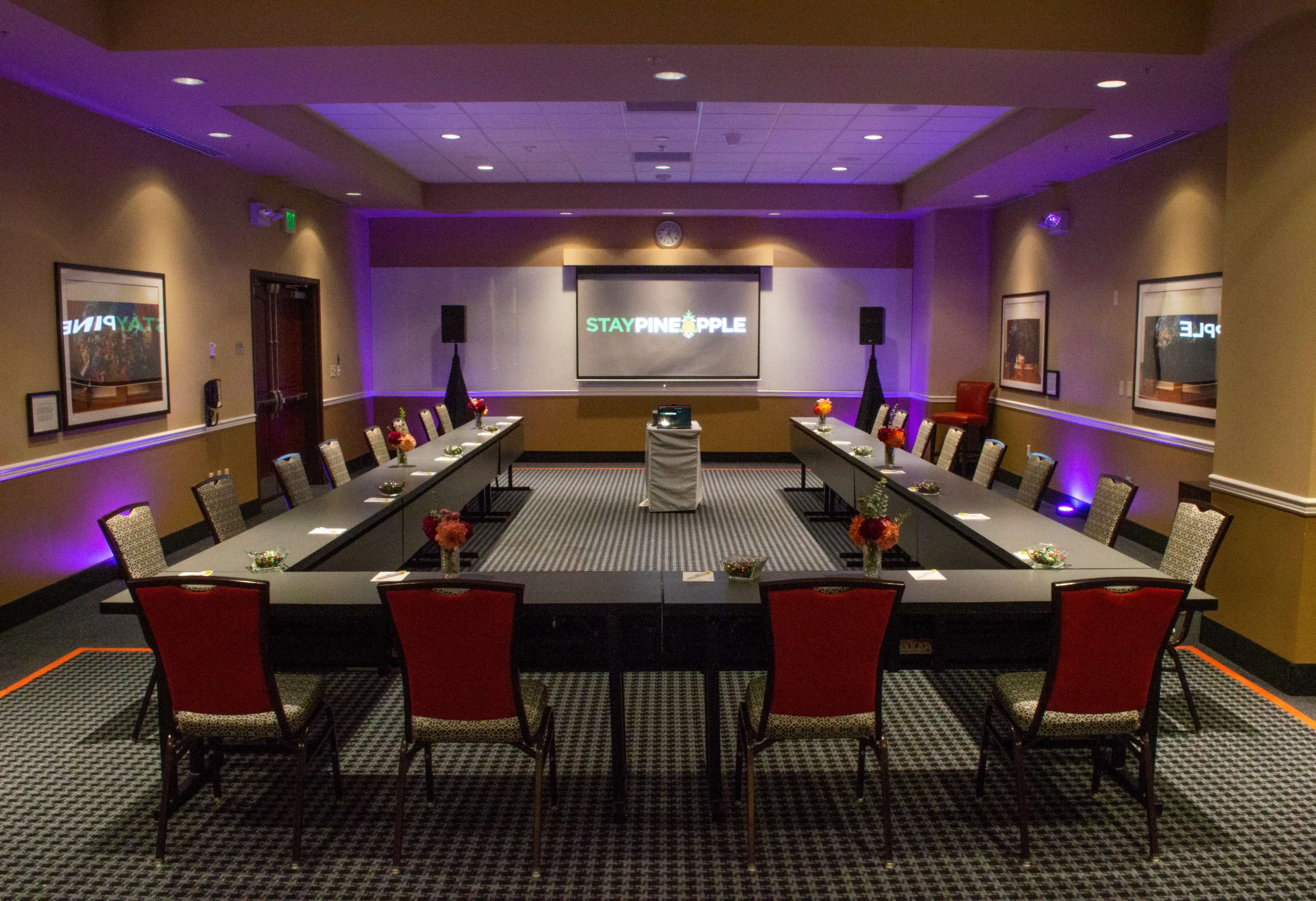 A conference room set up with two rows of tables arranged in a U-shape, decorated with chairs and floral centerpieces, and equipped with audio-visual equipment for presentations.