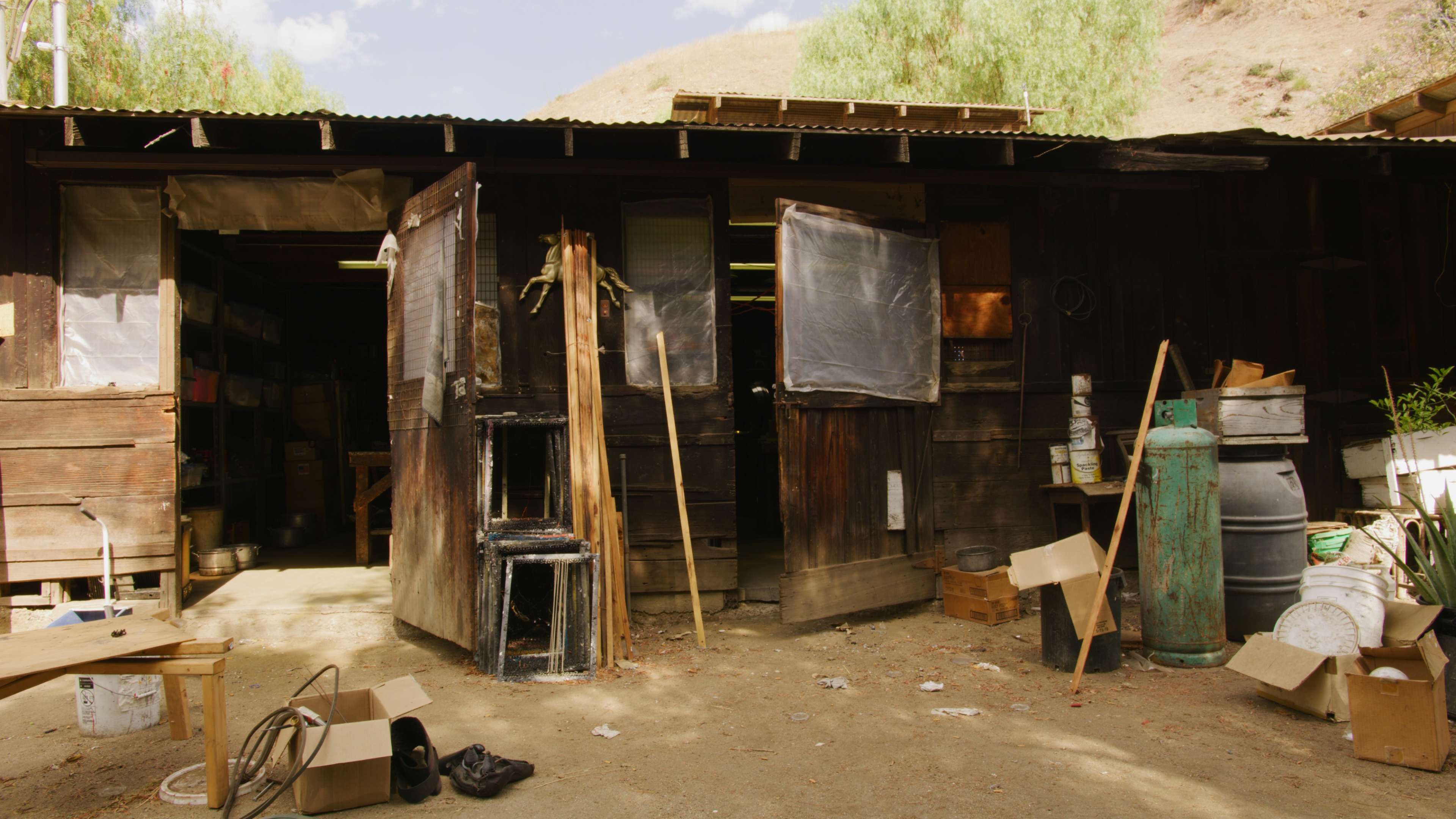 A rustic wooden shed with open doors, surrounded by scattered boxes and tools on a dirt floor.